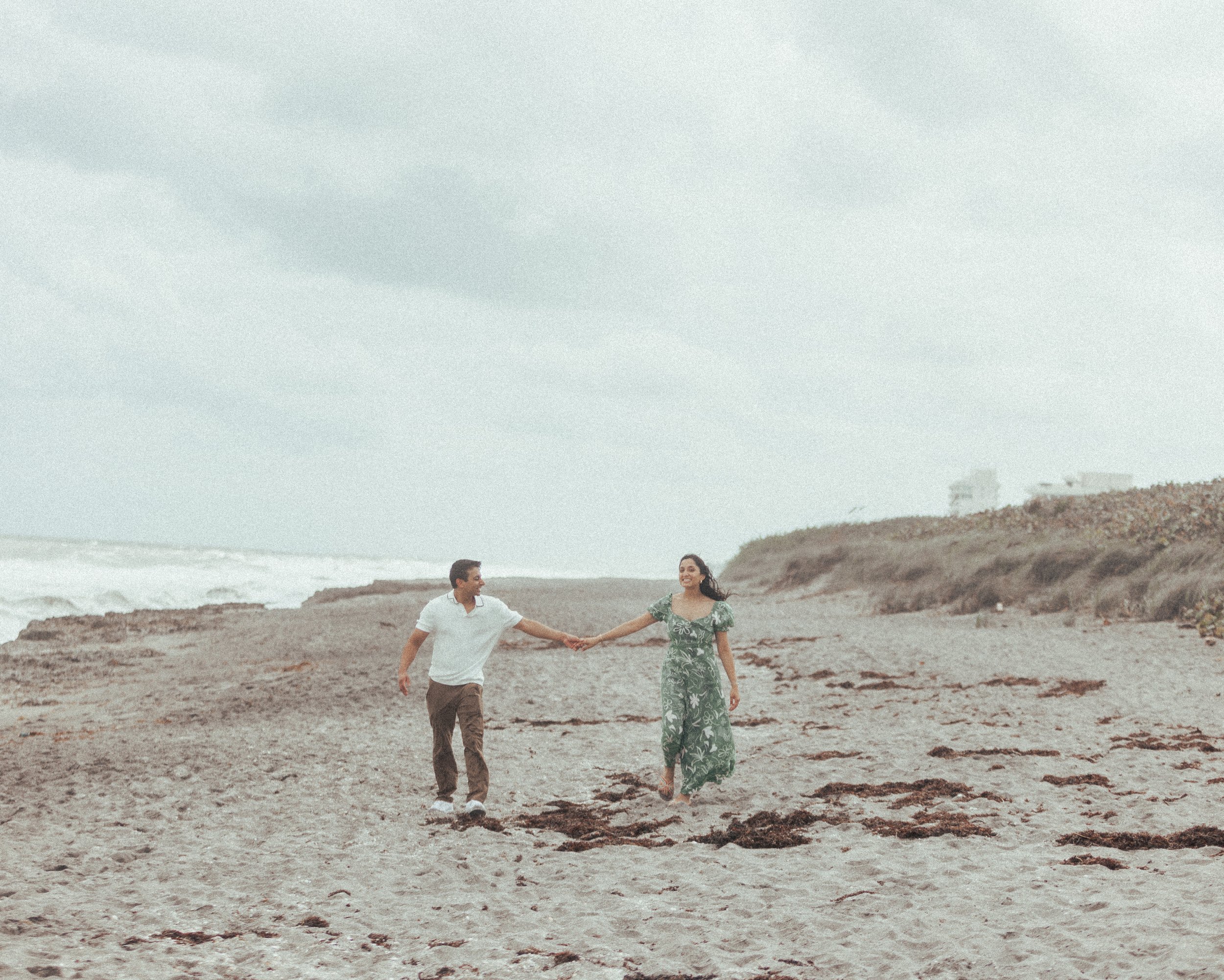 A couple walking hand in hand along a beach with cloudy sky and dunes in the background.