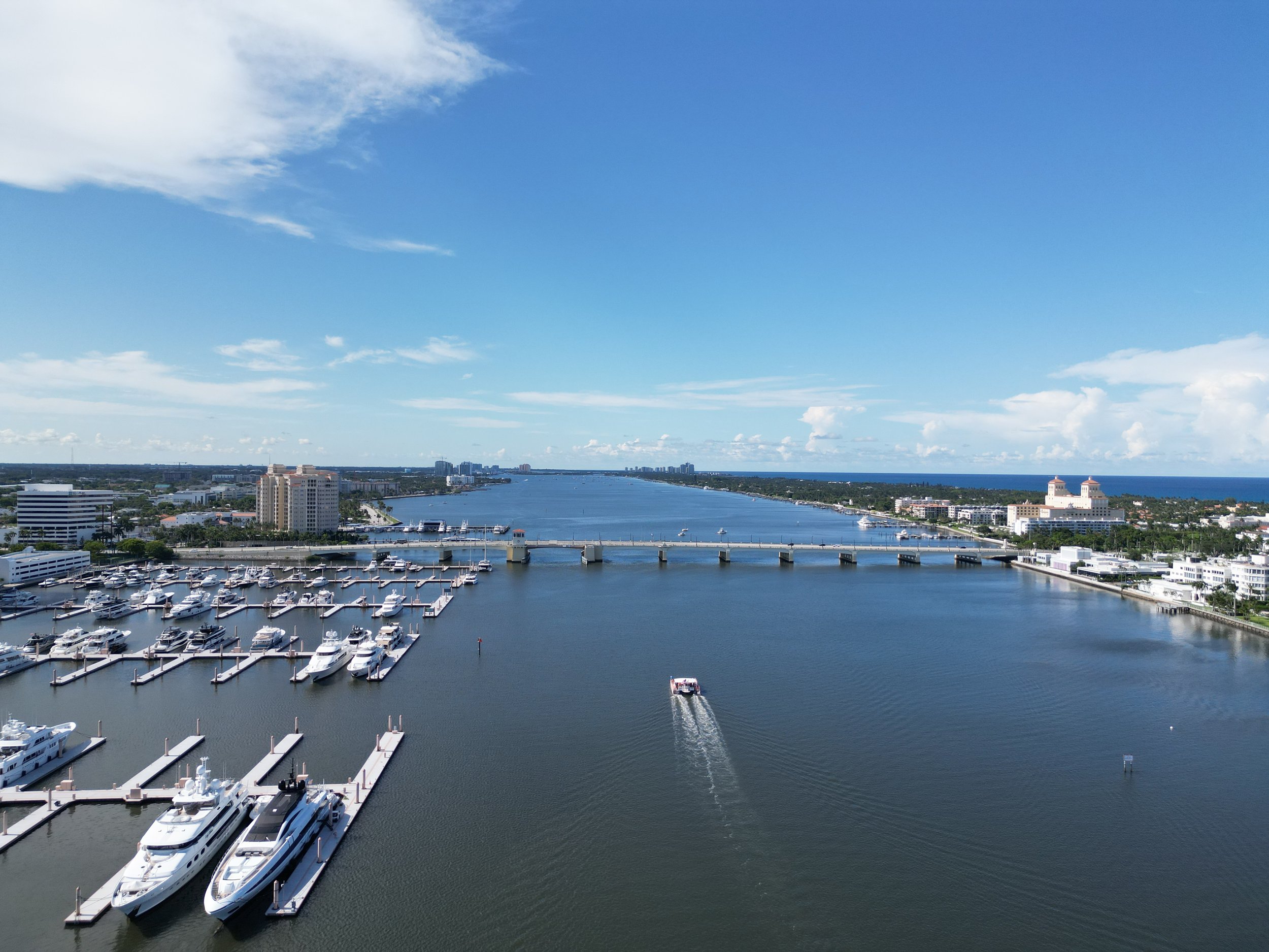 Aerial view of a marina with yachts, a bridge crossing the waterway, and urban buildings along the shoreline under a partly cloudy sky.