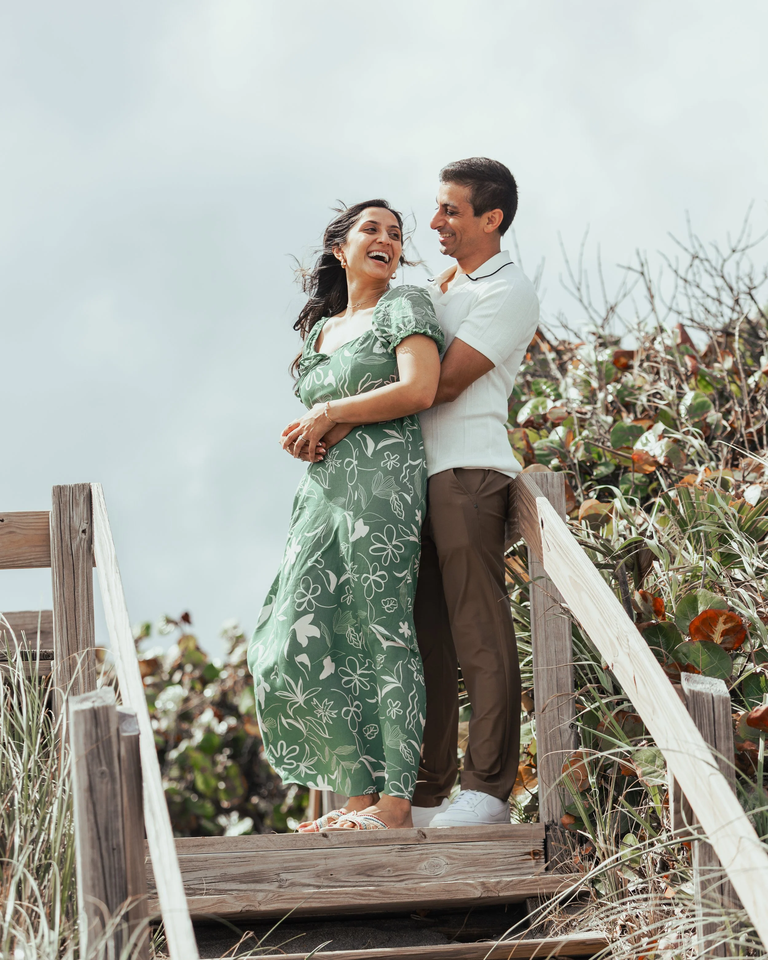 A happy couple standing on a wooden bridge outdoors, embracing and laughing with a cloudy sky in the background.