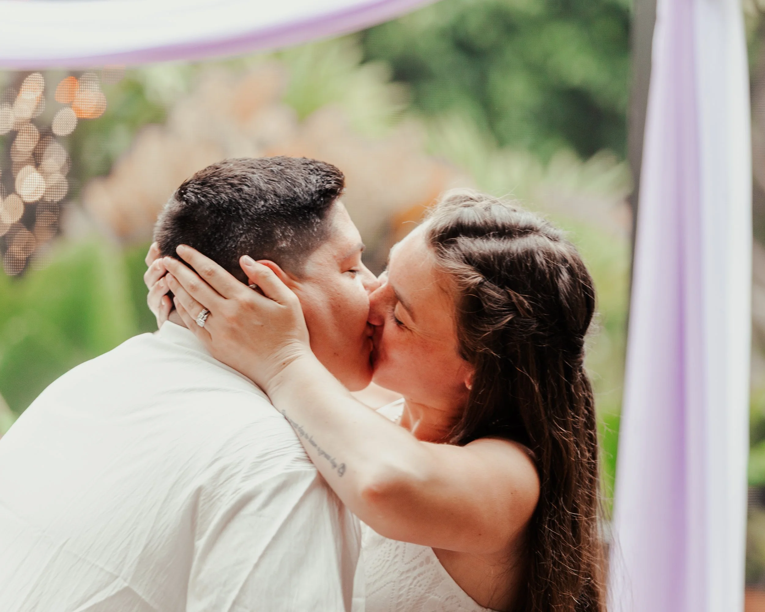 A couple kissing, holding each other's faces, outdoors with blurred greenery in the background.