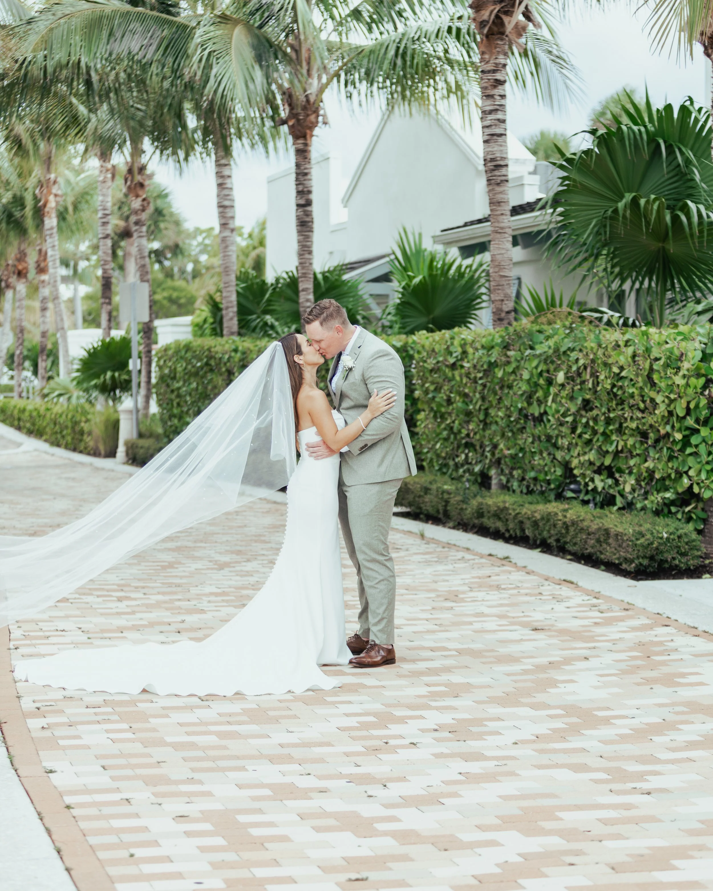 A newlywed couple sharing a kiss outdoors on a brick pathway with palm trees and greenery in the background.