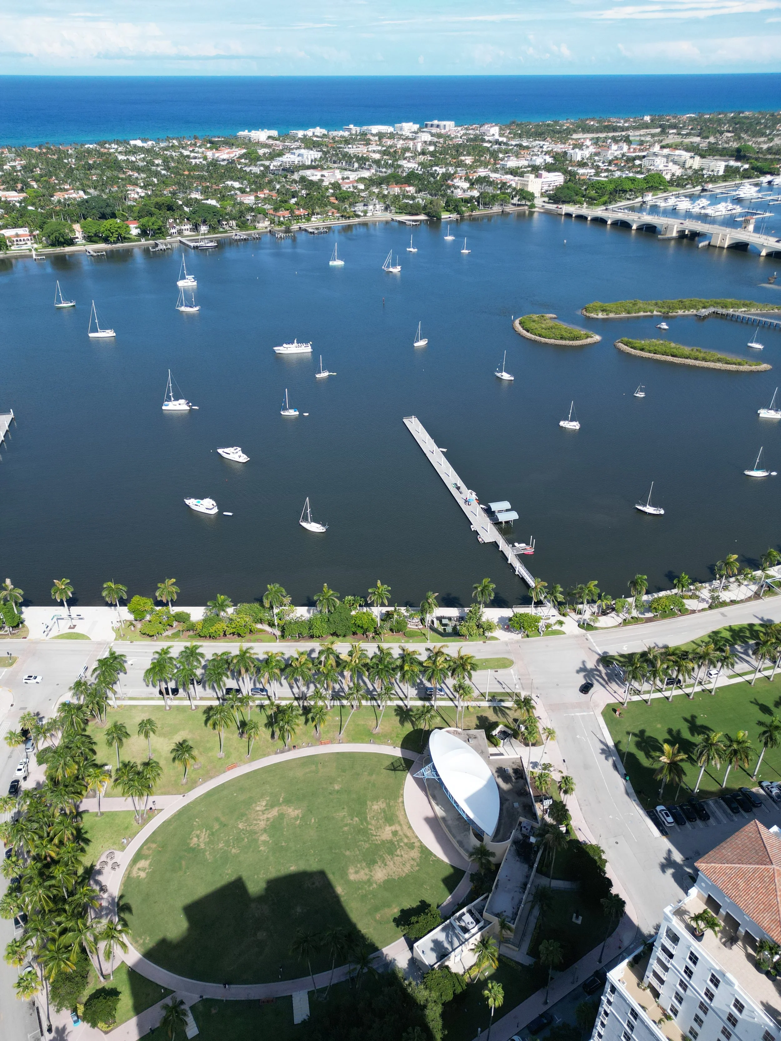 Aerial view of a coastal city with a marina, boats, green park, and ocean in the background.