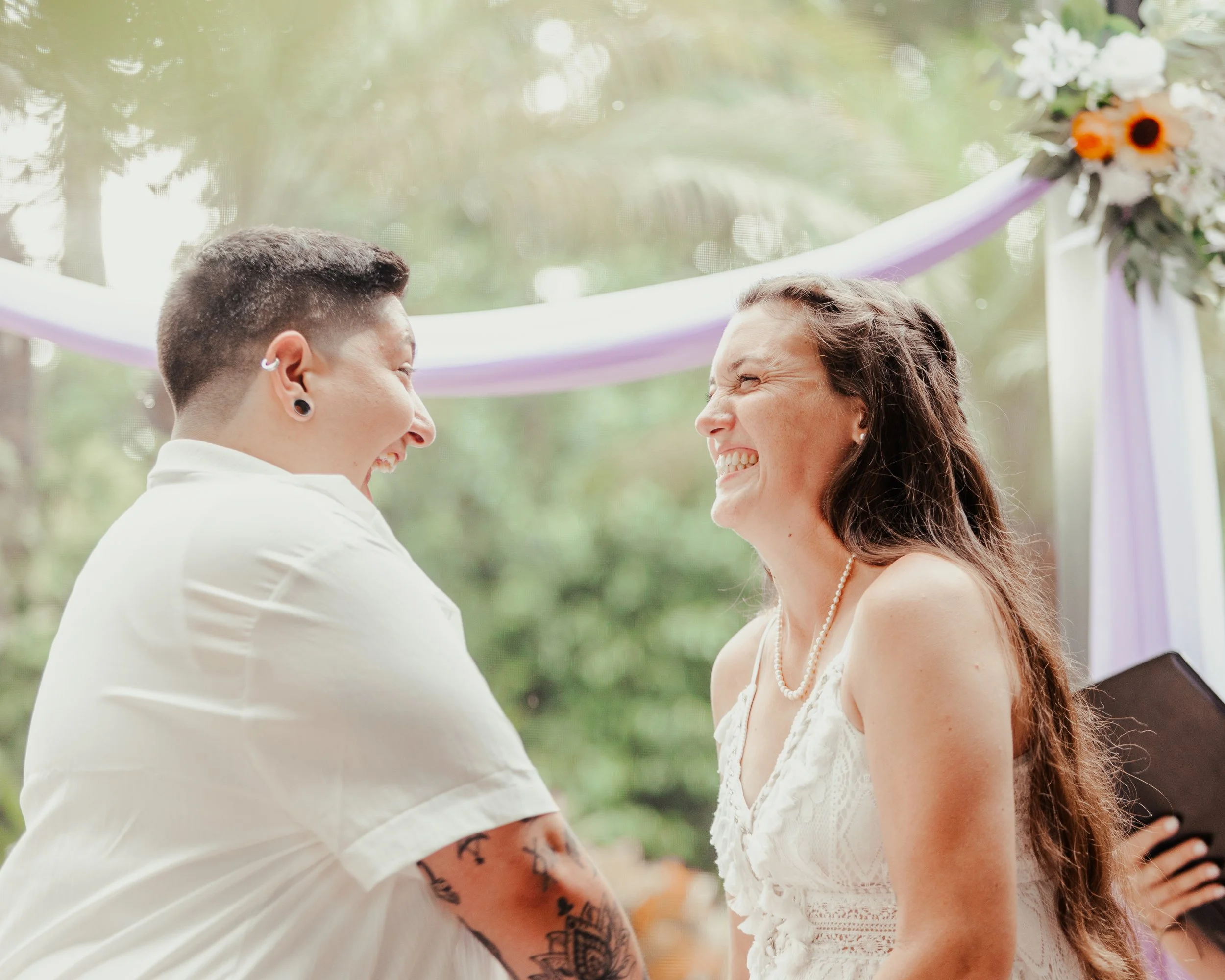 Two women smiling at each other during a wedding ceremony, standing under a decorated arch with greenery and flowers.