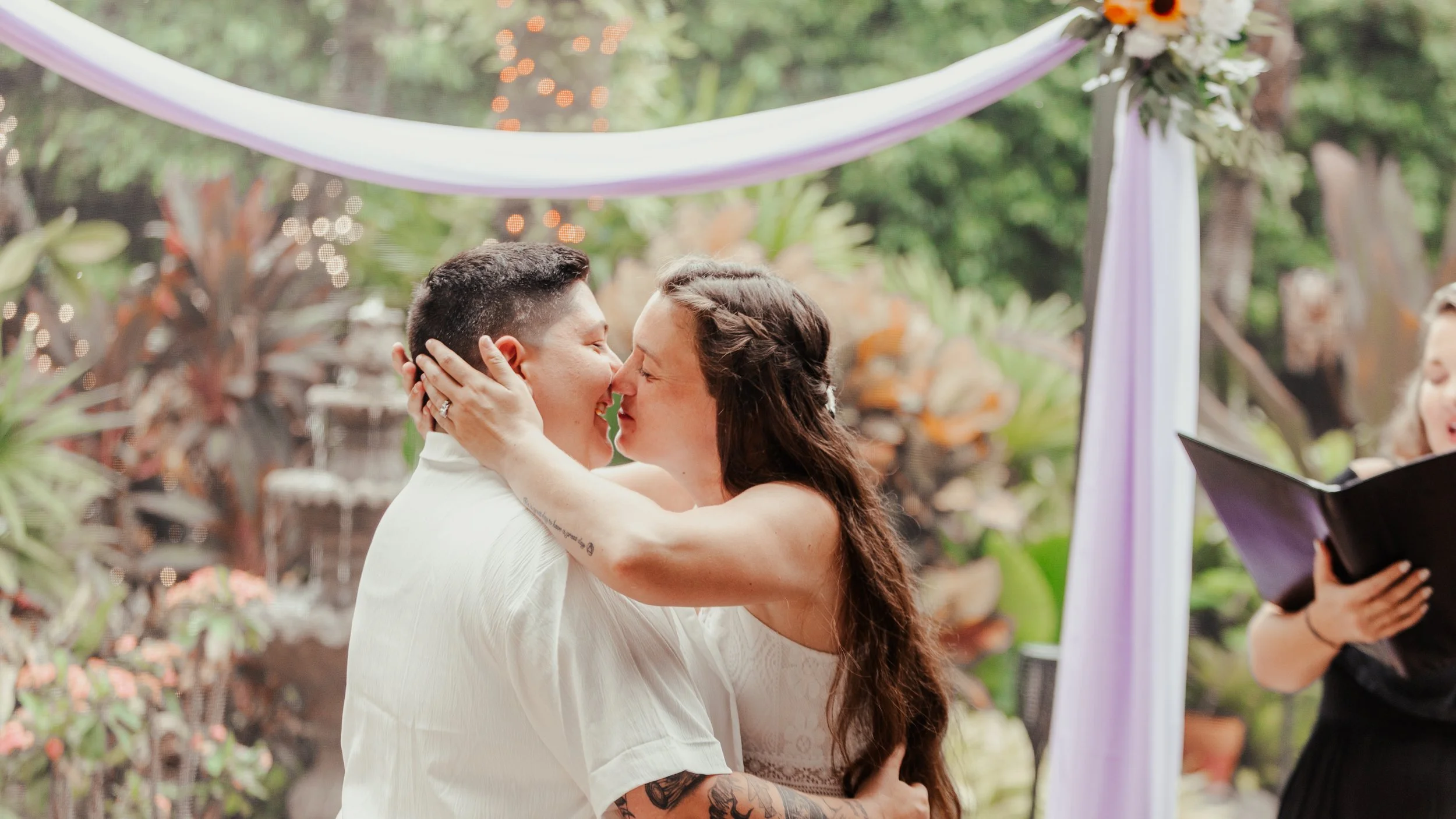 A couple is sharing a kiss during a wedding ceremony outdoors, with lush greenery and plants in the background, and an officiant holding a black book on the right side.