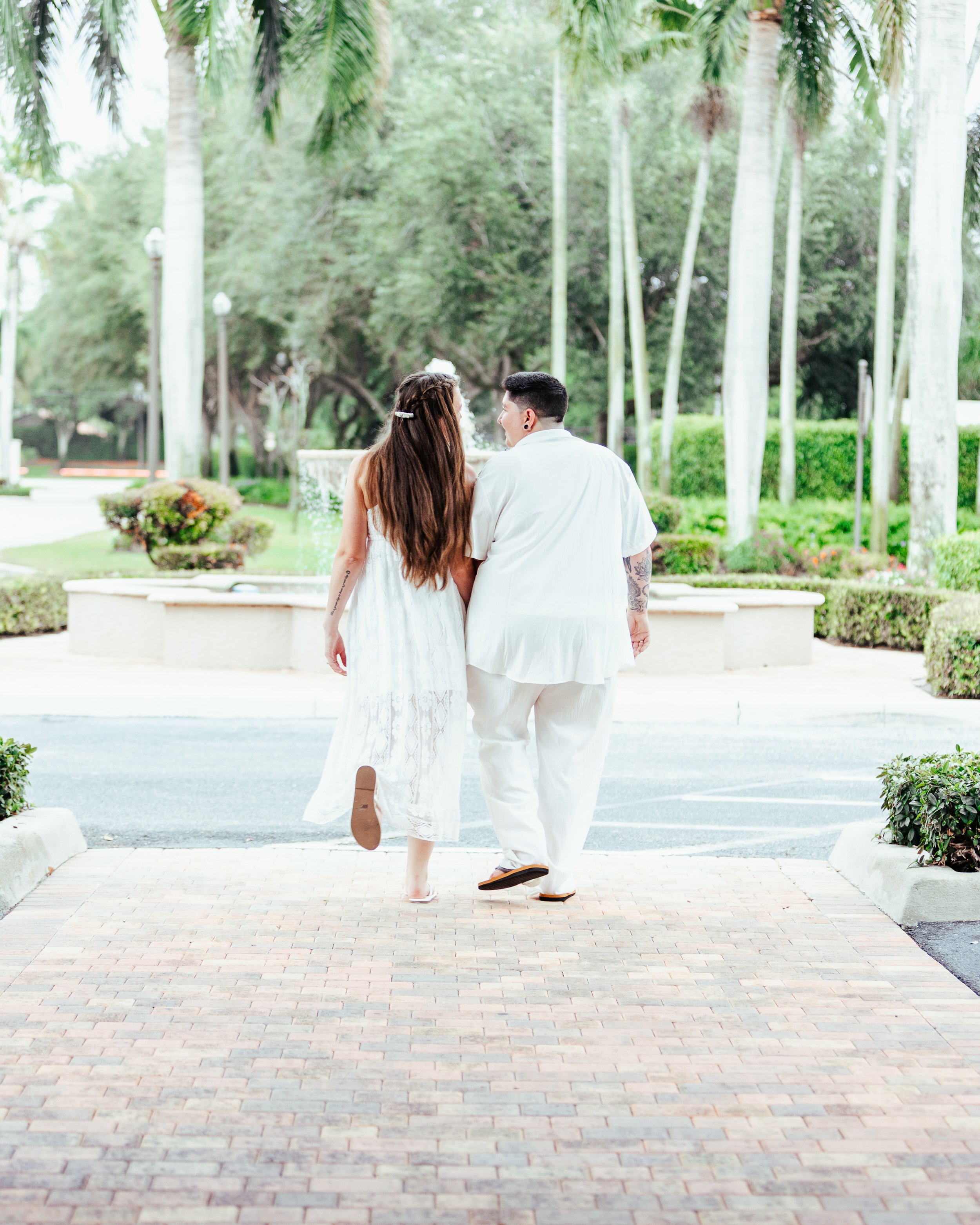 A couple dressed in white walking hand in hand in a park with trees and greenery.