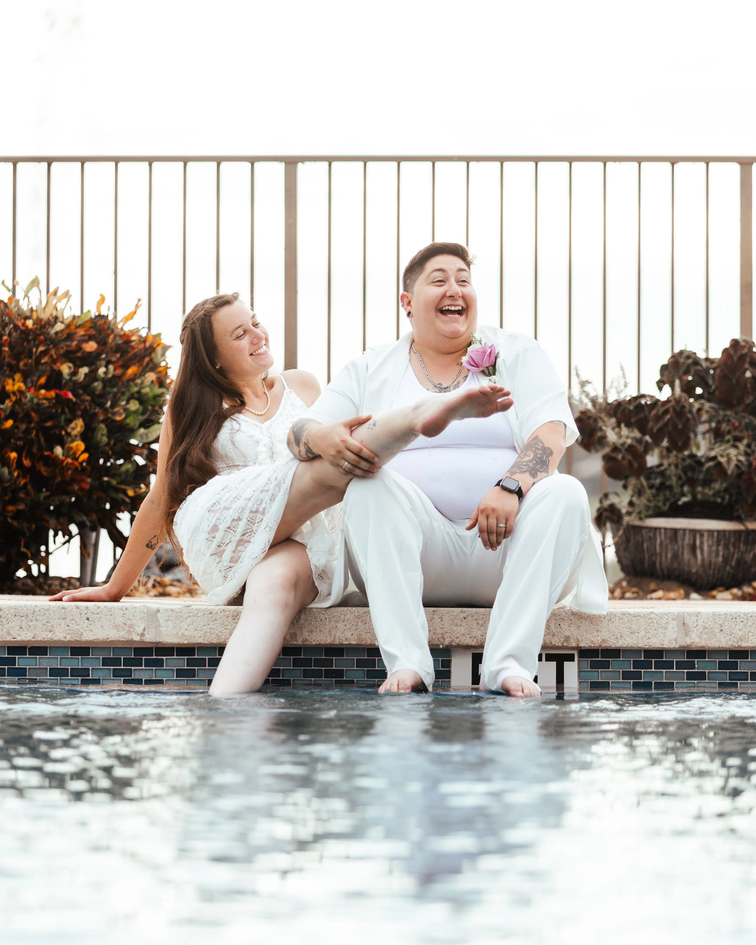 A couple in white clothing celebrating by a pool, with the man smiling and holding the woman's leg, while the woman is smiling and leaning on him, surrounded by plants and a fence.