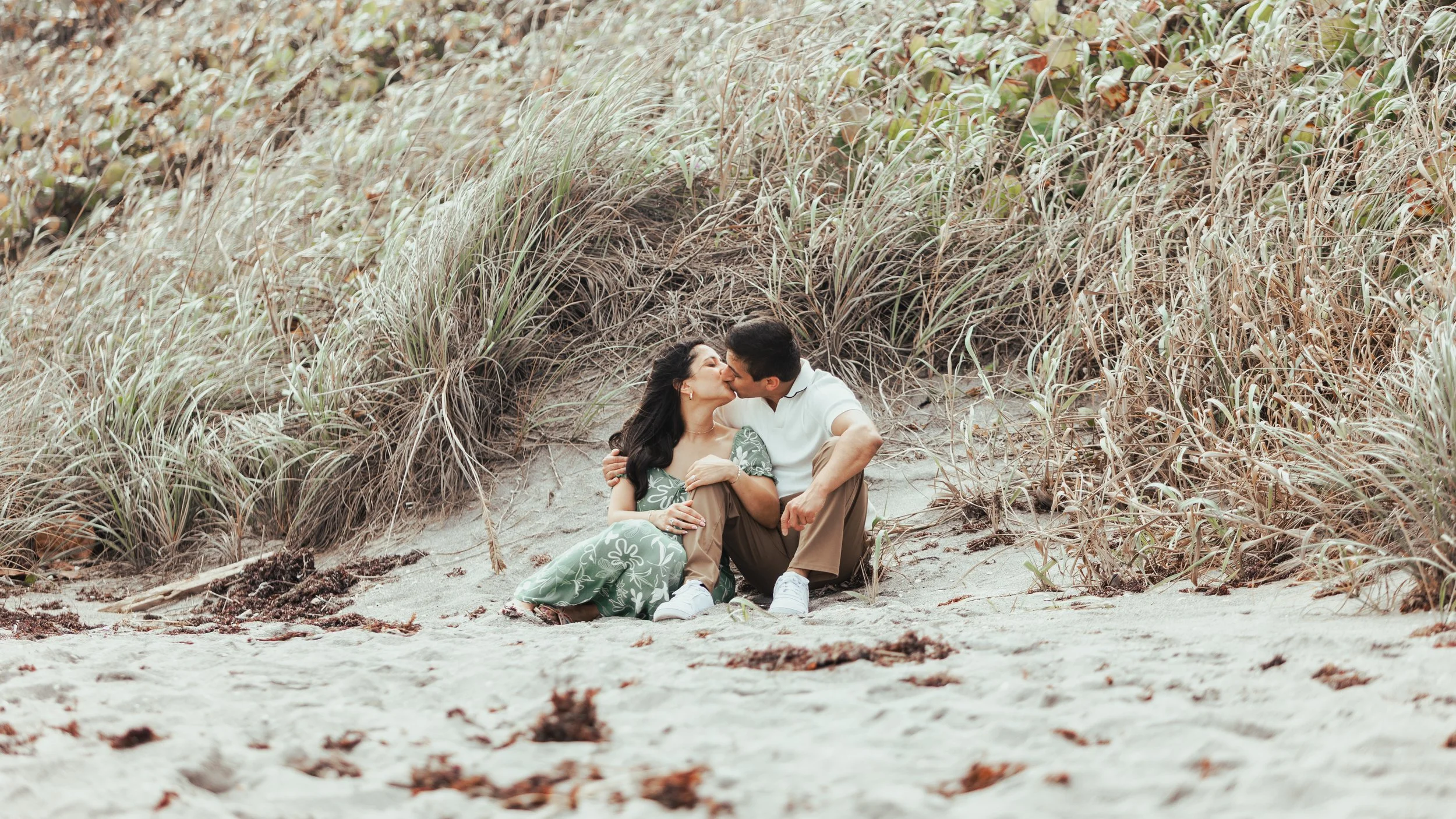 A couple sitting on the sandy beach, kissing, surrounded by tall grass and dunes.