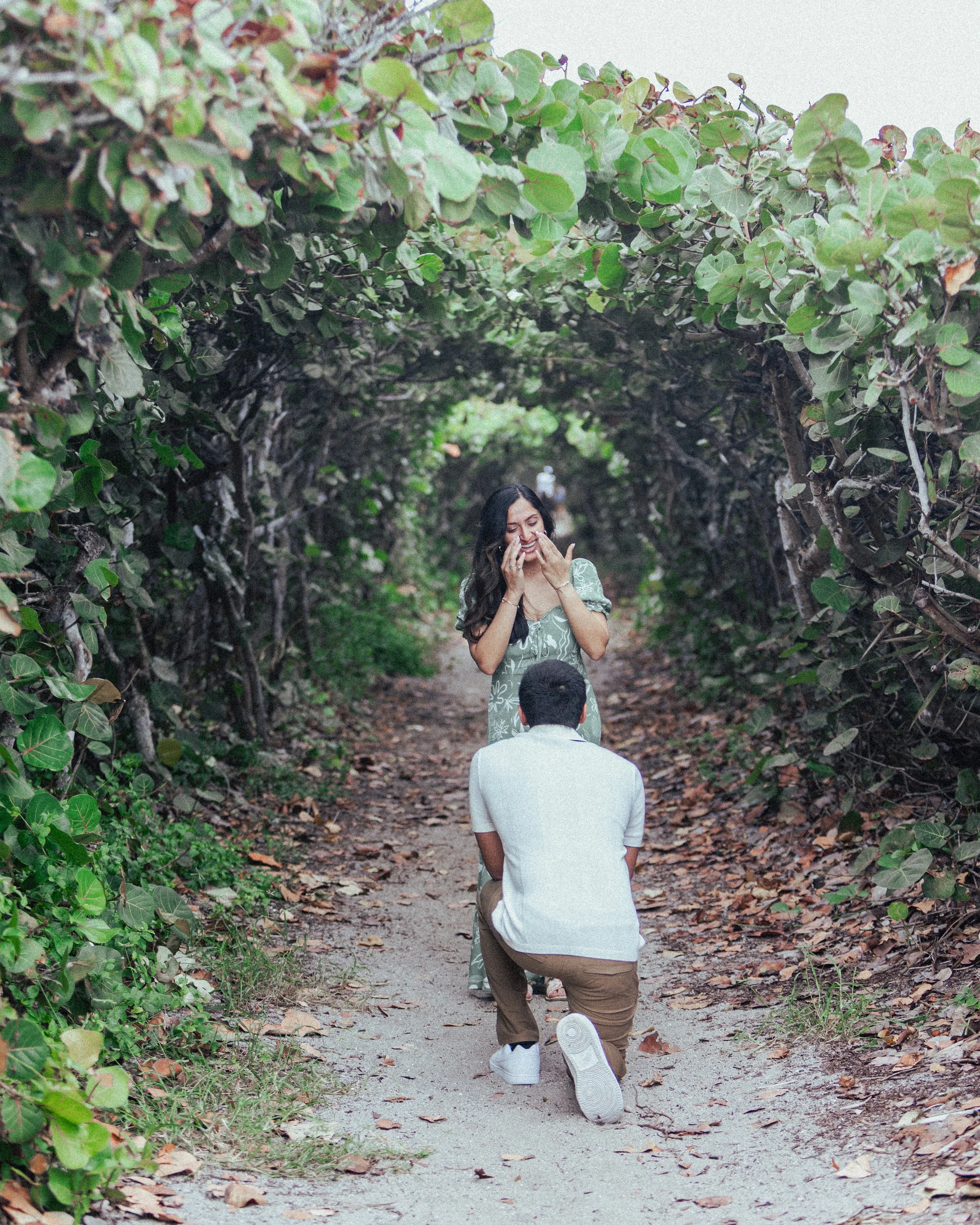 A woman is proposing to a man in a leafy tunnel on a dirt path, with the woman smiling and covering her mouth, and the man kneeling with his back to the camera.