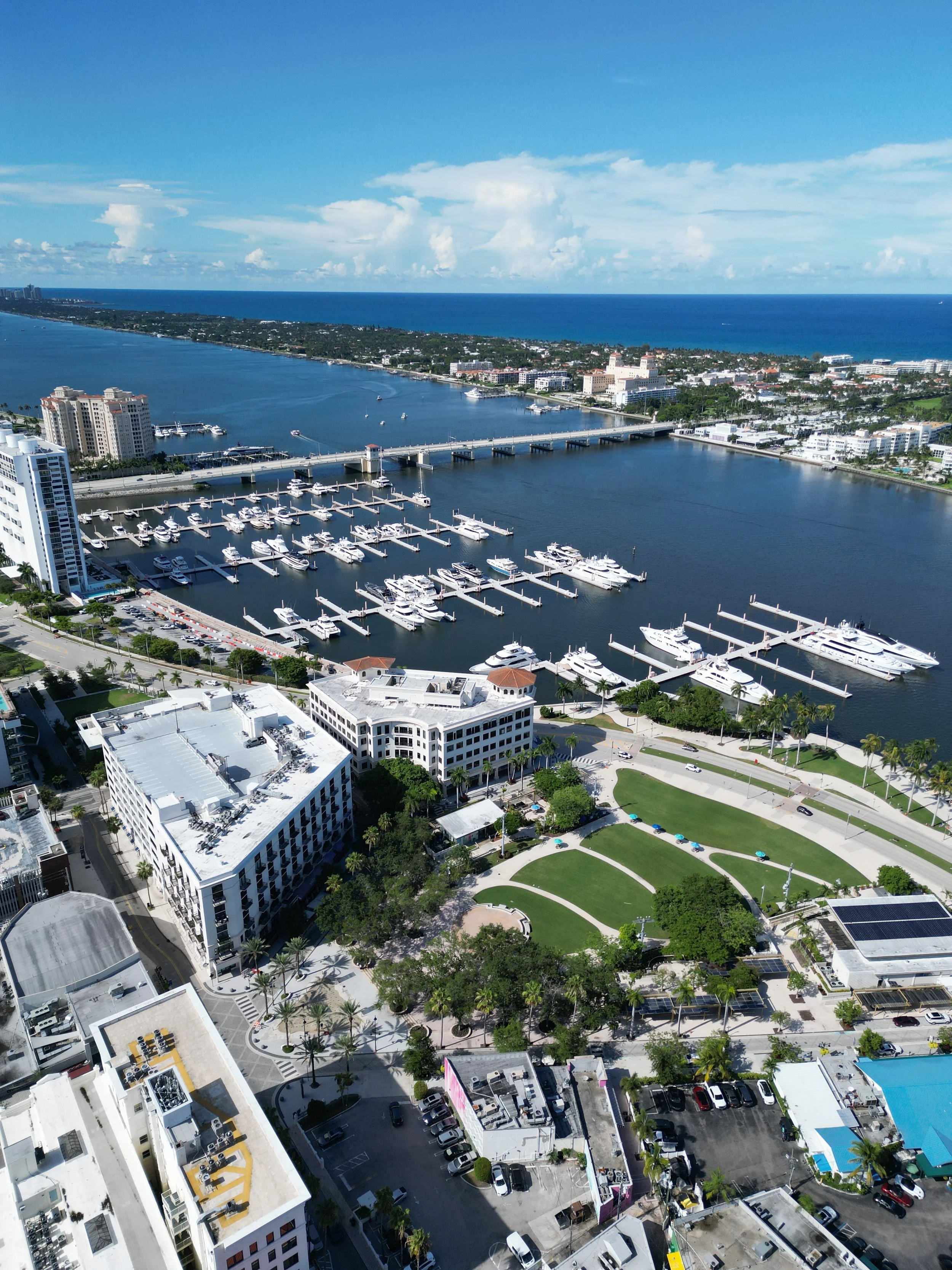 Aerial view of a marina with boats docked, high-rise buildings, green park area, and waterway under a partly cloudy sky.
