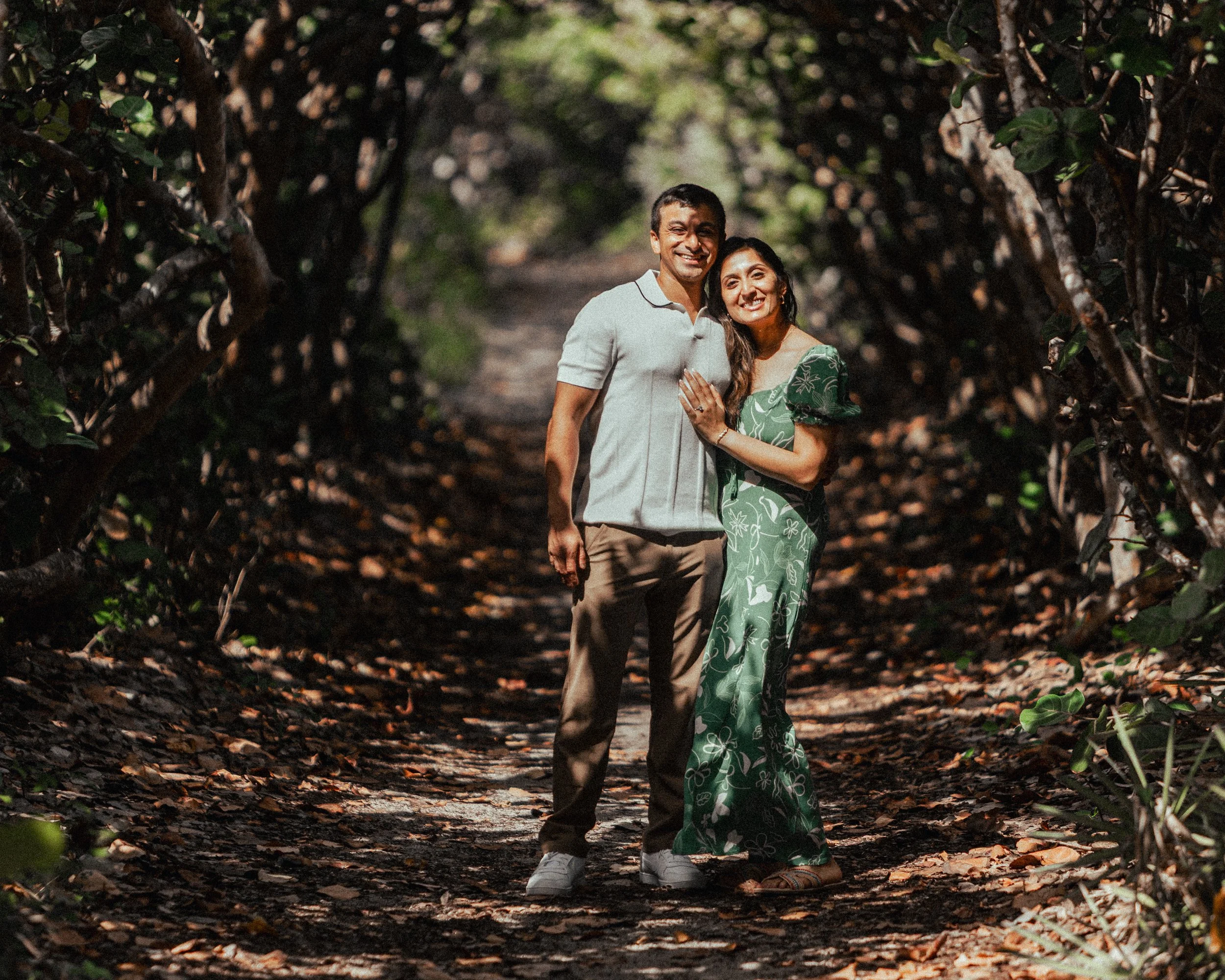 A smiling couple stands close together on a forest path, surrounded by trees and foliage.
