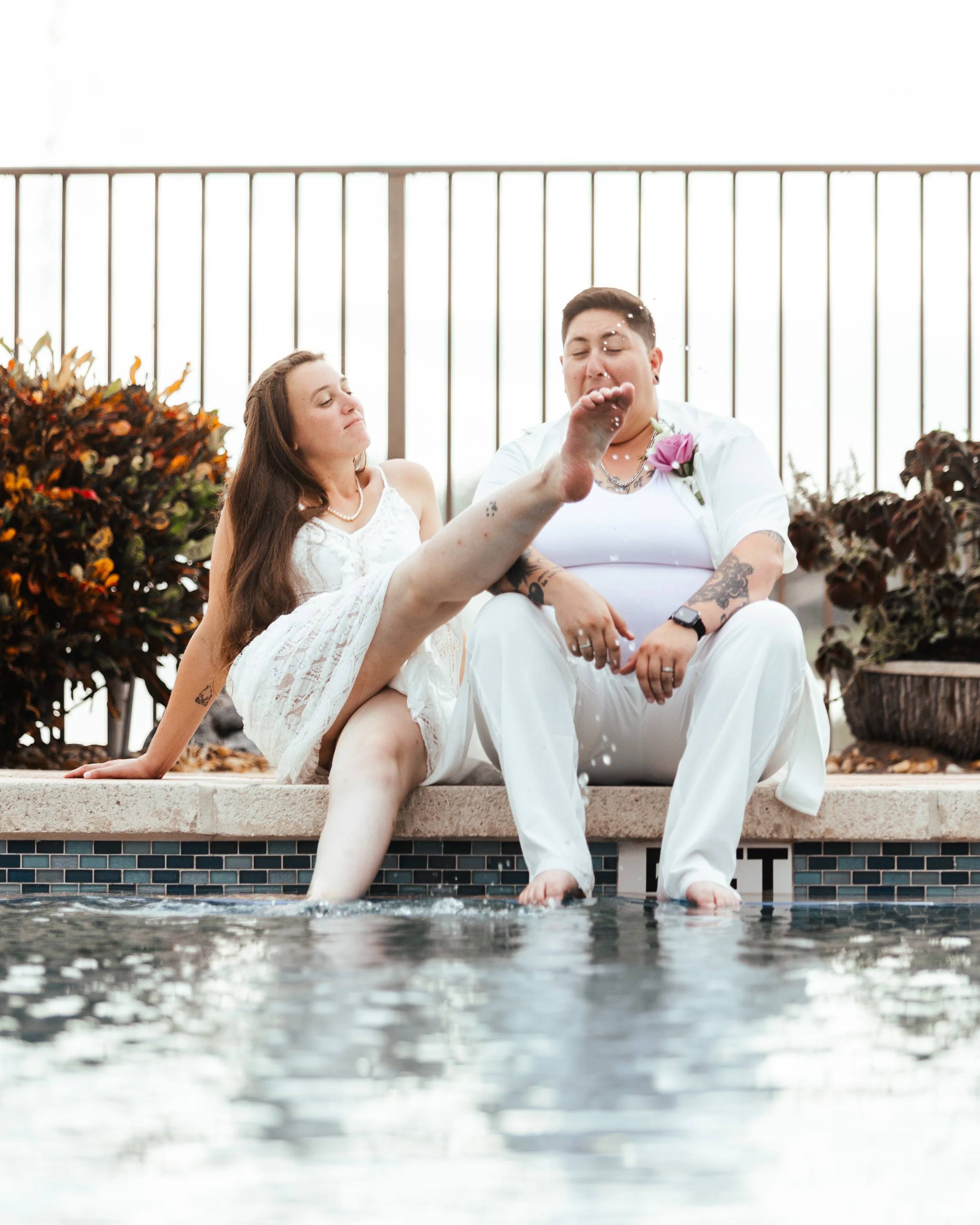 Two people sitting by a pool, one splashing water on the other during a casual outdoor event.