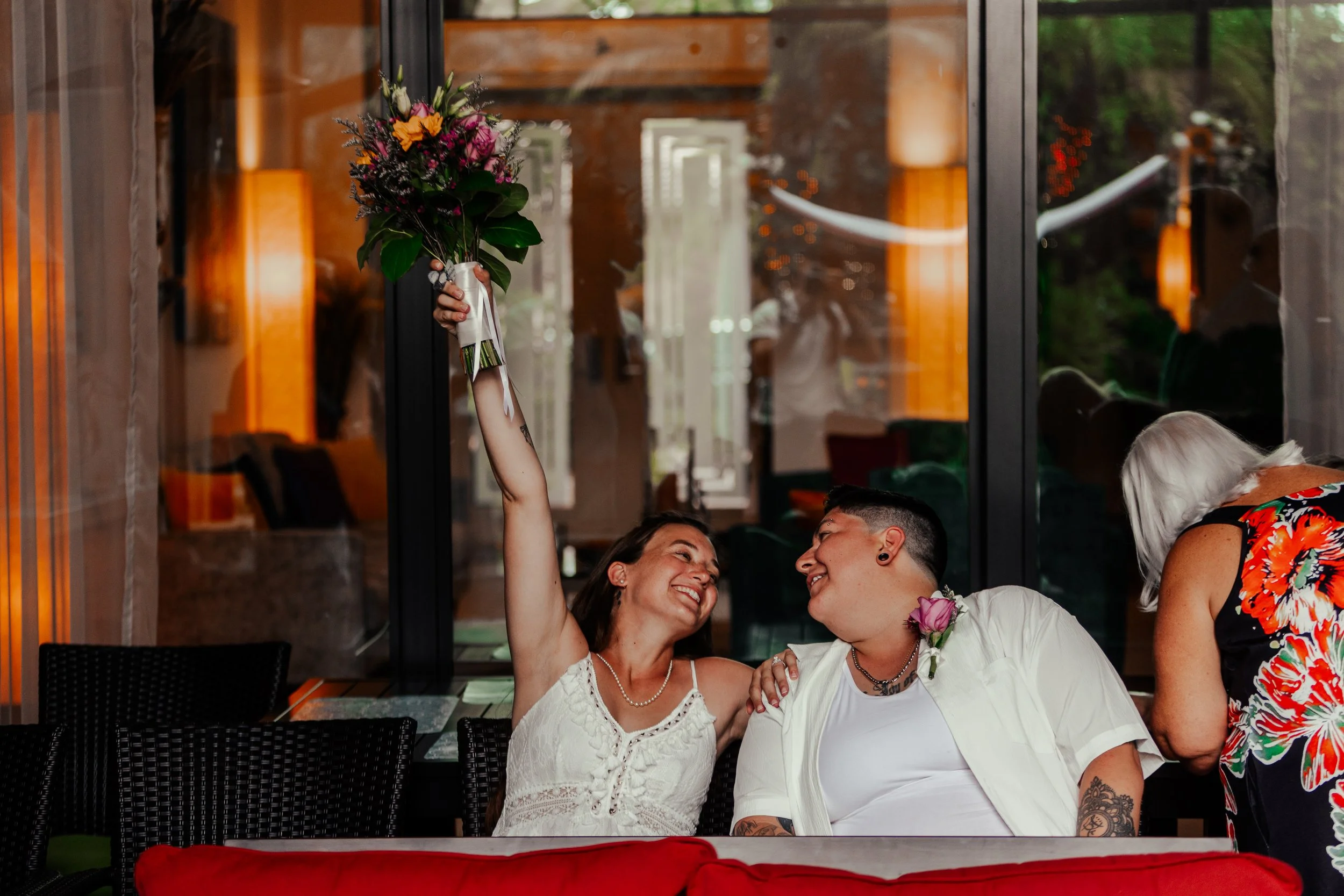 A woman holding up a bouquet of flowers, smiling at a man, at a celebration or gathering inside a restaurant or cafe.