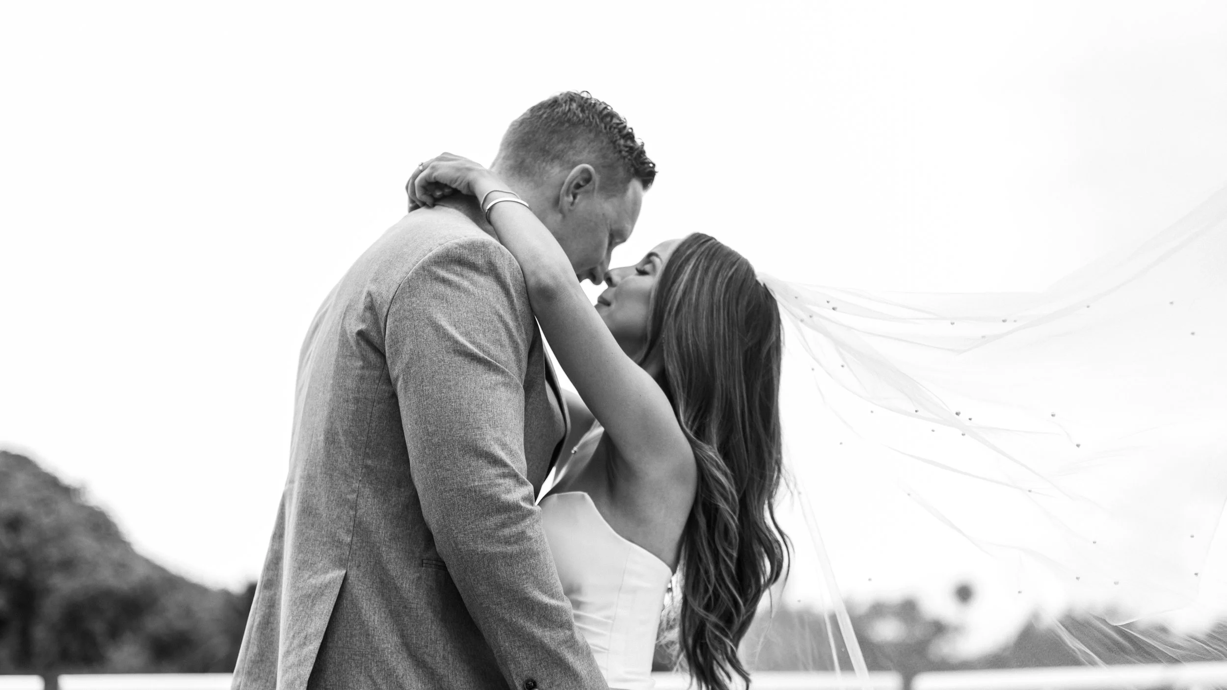 Black and white photo of a bride and groom embracing closely, their faces near each other, outdoors, with the bride's veil flowing in the wind.