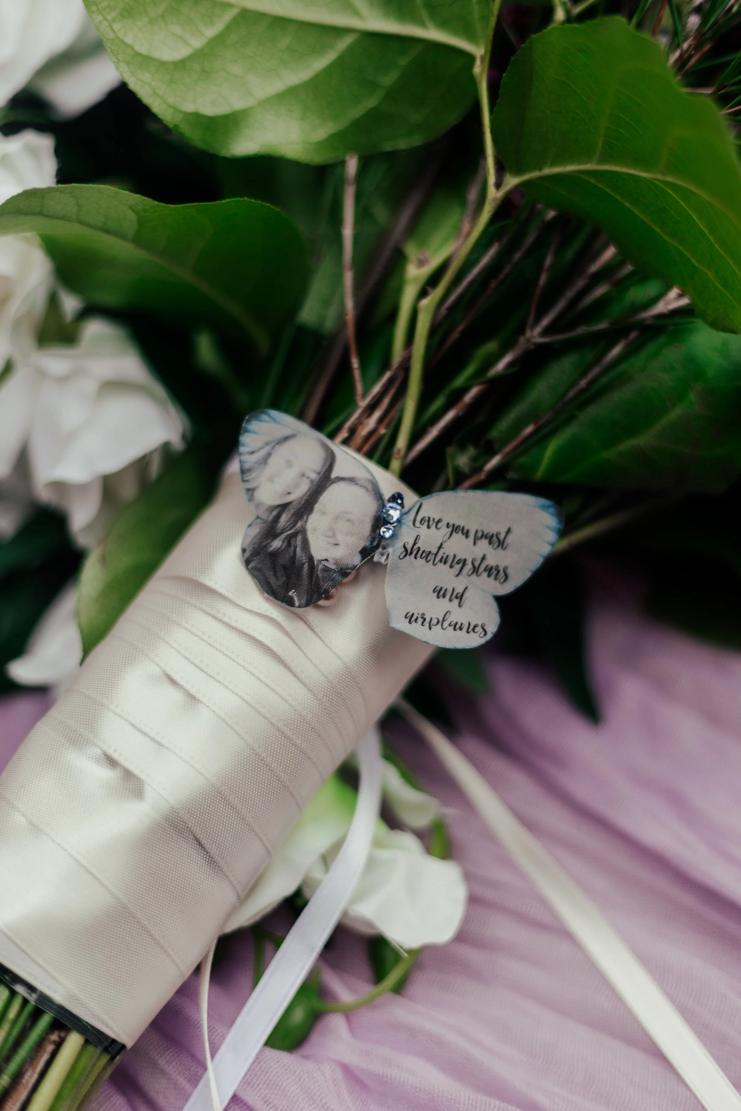 A bouquet of green leaves and white flowers with a ribbon-wrapped handle, with a butterfly-shaped decoration featuring a black-and-white photo of a person and a quote about love and shooting stars.
