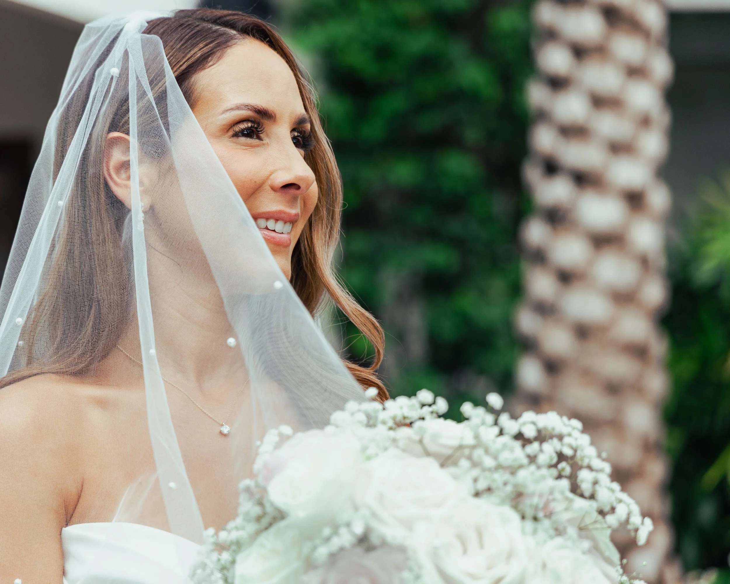 A smiling bride with long, wavy brown hair, wearing a veil and holding a bouquet of white flowers, outdoors with palm trees in the background.