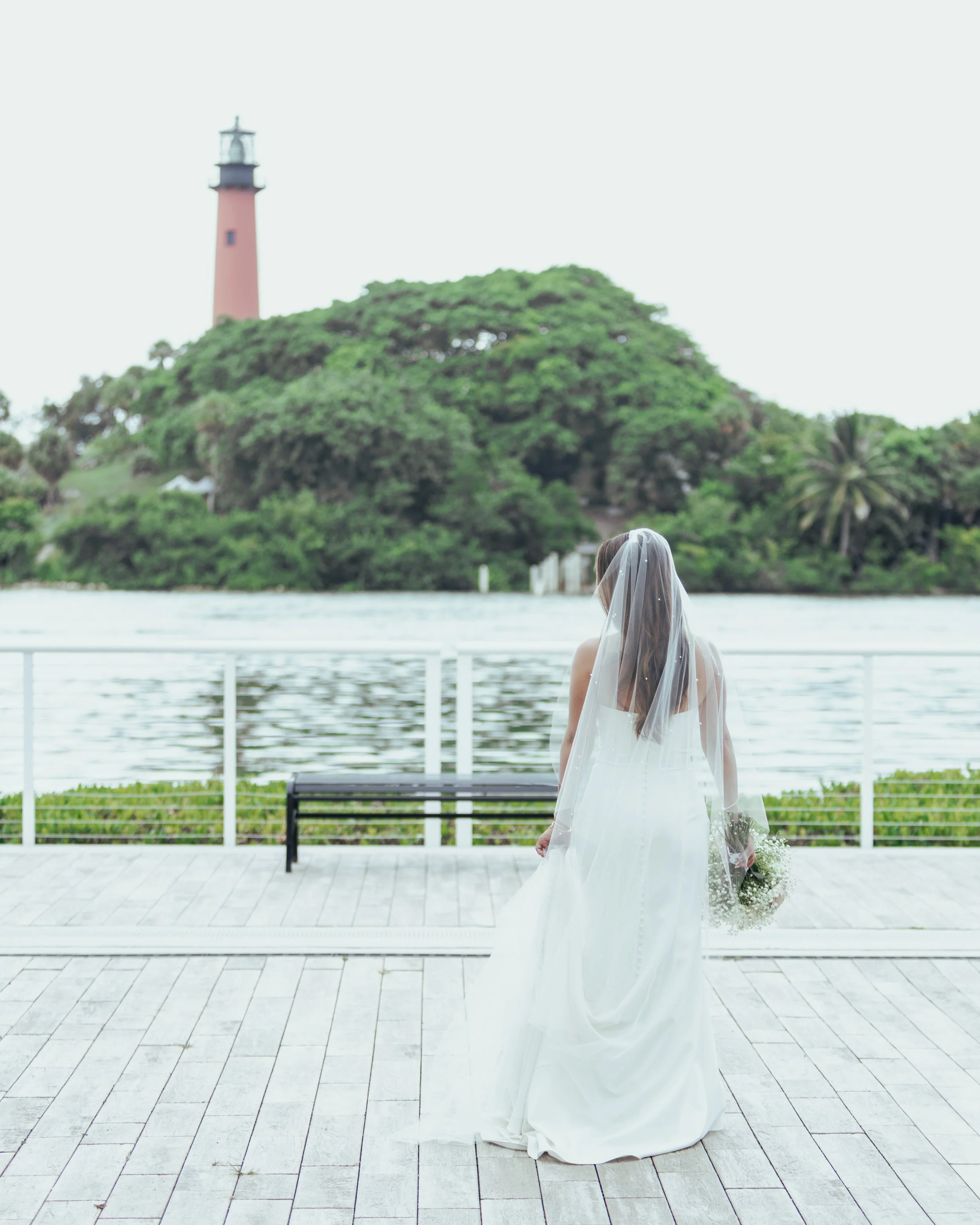 Bride in a white wedding dress and veil holding a bouquet, standing on a dock by water with green trees and a lighthouse in the background.