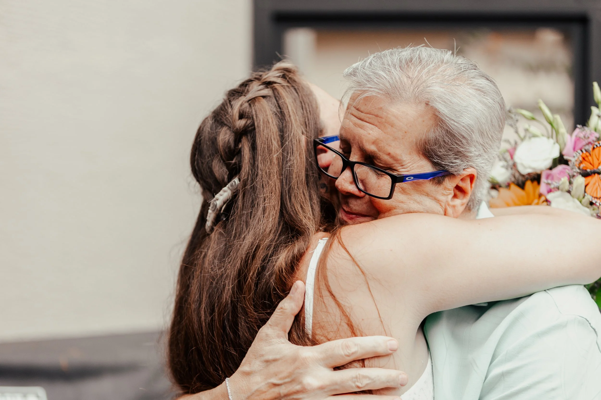 A young girl with braided hair hugging an elderly woman with gray hair and glasses, both smiling in a warm embrace, with flowers in the background.