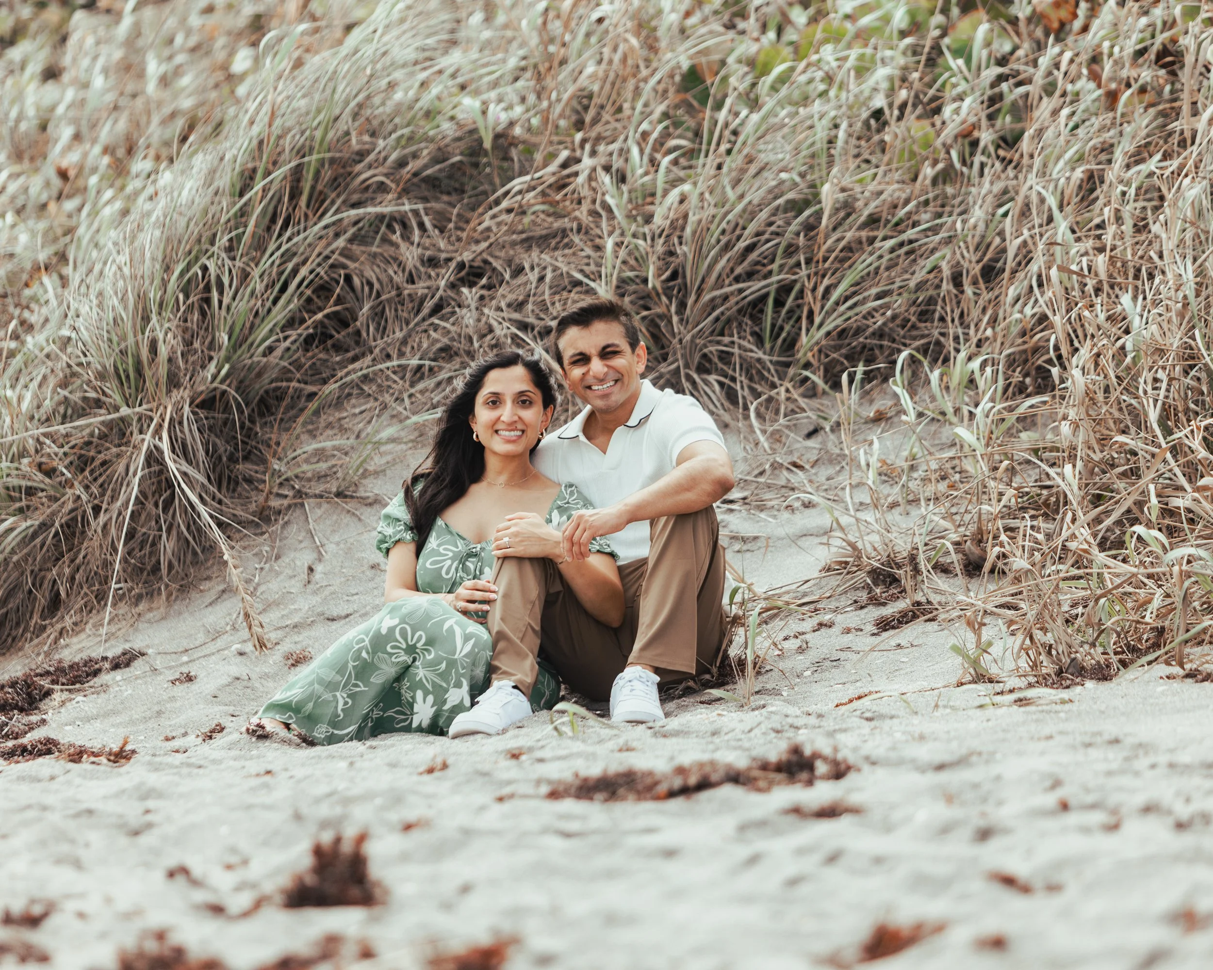 A couple sitting close together on the sandy beach with dune grass behind them. They are smiling at the camera, the woman wearing a floral dress and the man in a white shirt and tan pants.