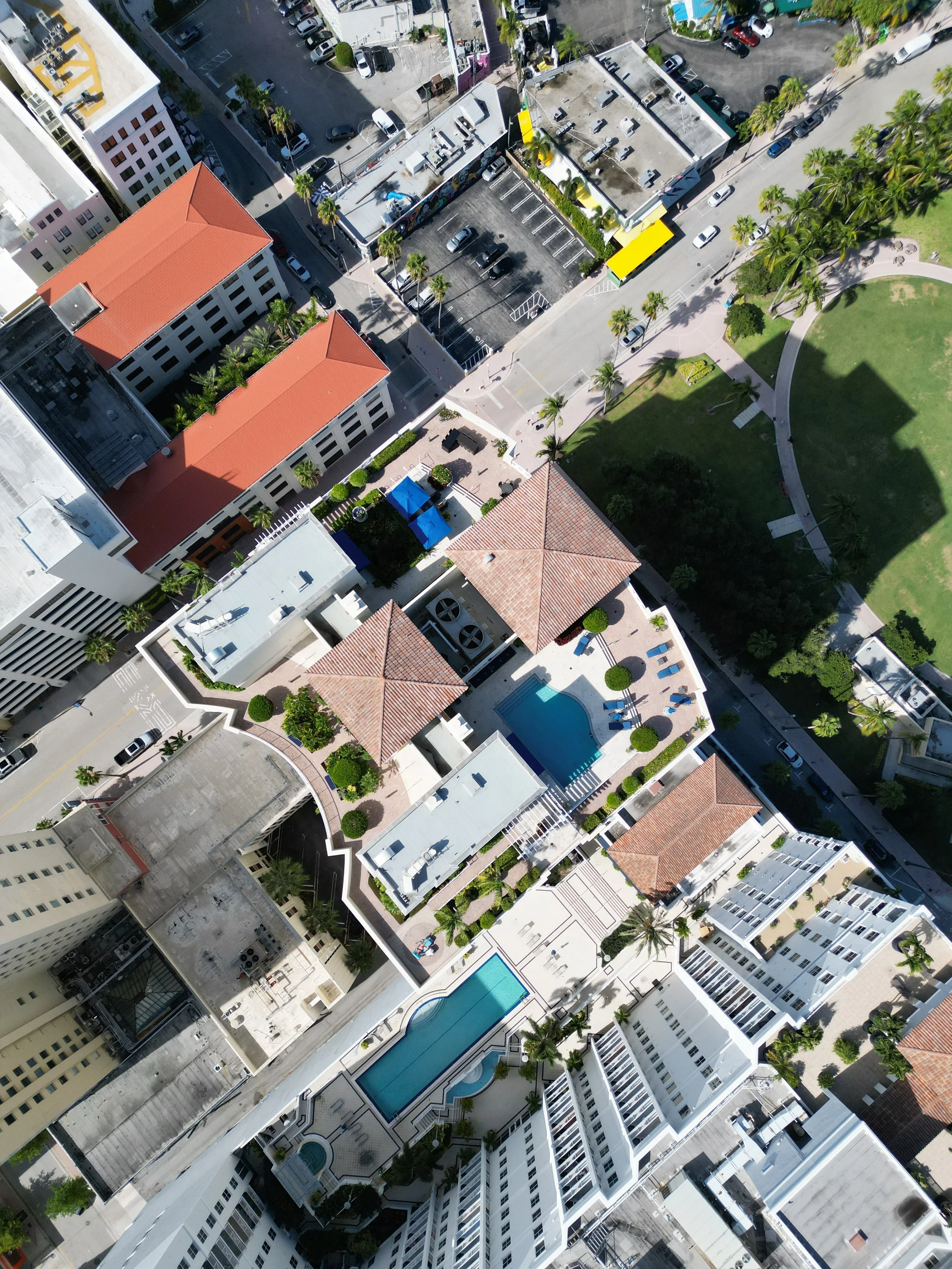An aerial view of a city block featuring a residential complex with two pools, surrounded by buildings, green spaces, and parking lots.