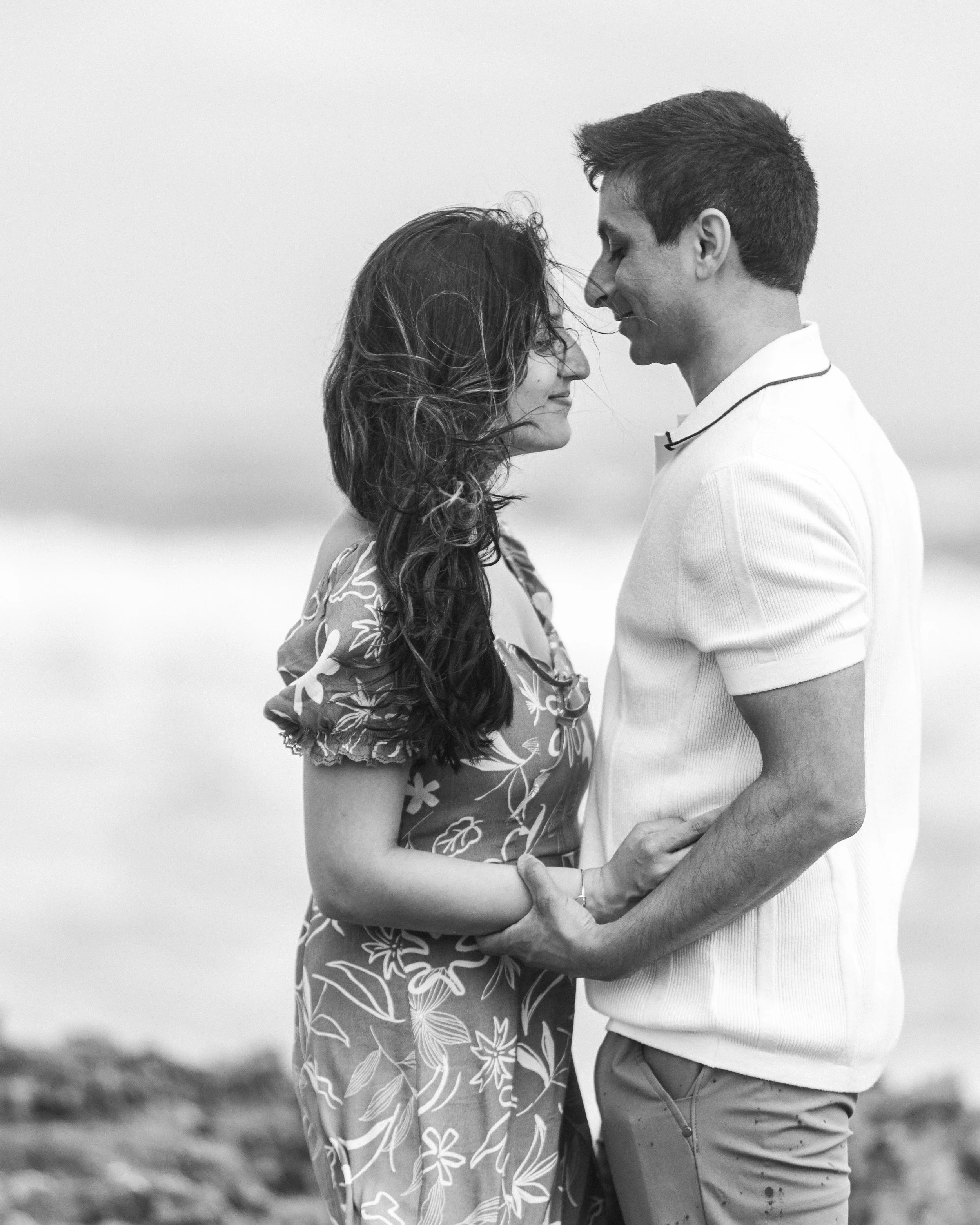 A black and white photo of a man and woman facing each other on a beach, holding hands and about to kiss.