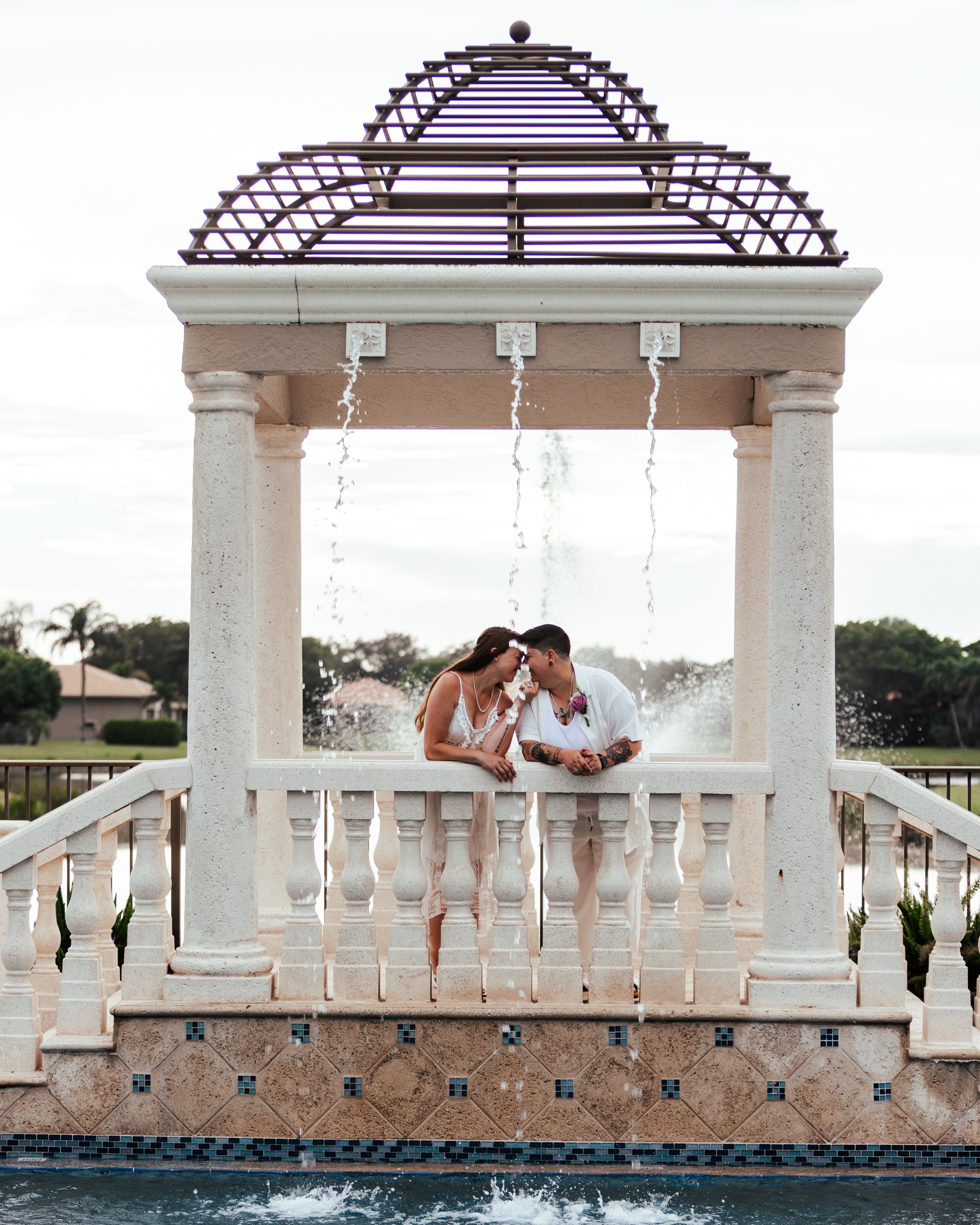 A couple is leaning on a white balustrade under a decorative fountain with water flowing from the top. They are touching foreheads and smiling at each other. The fountain is part of an ornate outdoor structure with columns and a tiered roof, set in a