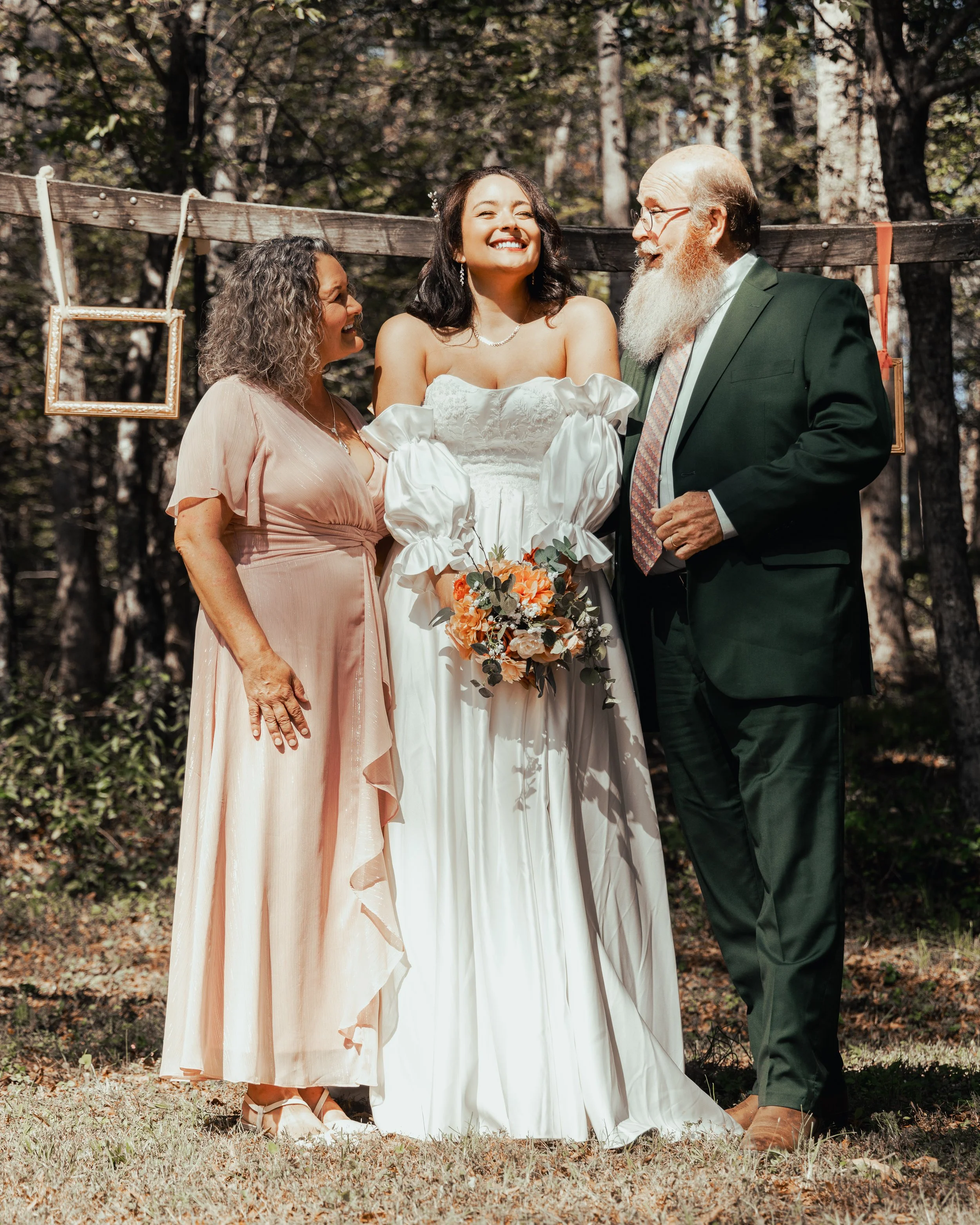 A bride standing outdoors with her parents during a wedding ceremony, holding a bouquet of flowers, surrounded by trees.