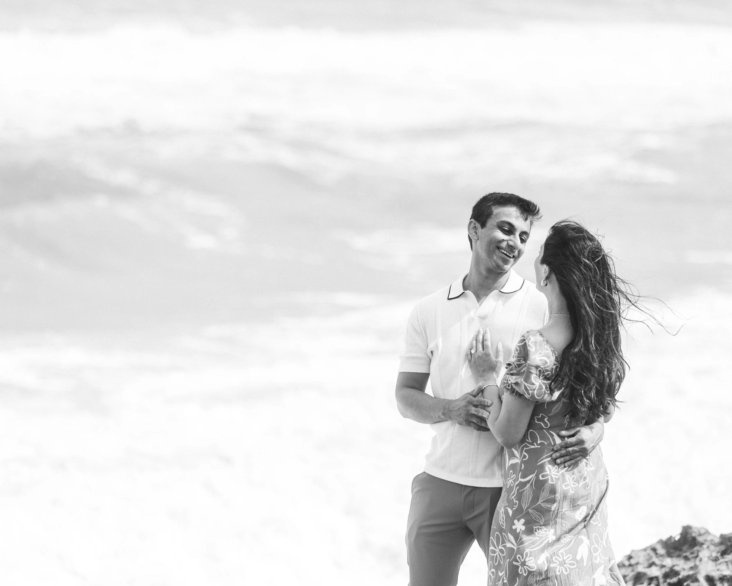 A black and white photo of a man and woman standing outdoors, smiling at each other, with the man gently holding the woman's waist. The background features a cloudy sky and some rocks in the distance.