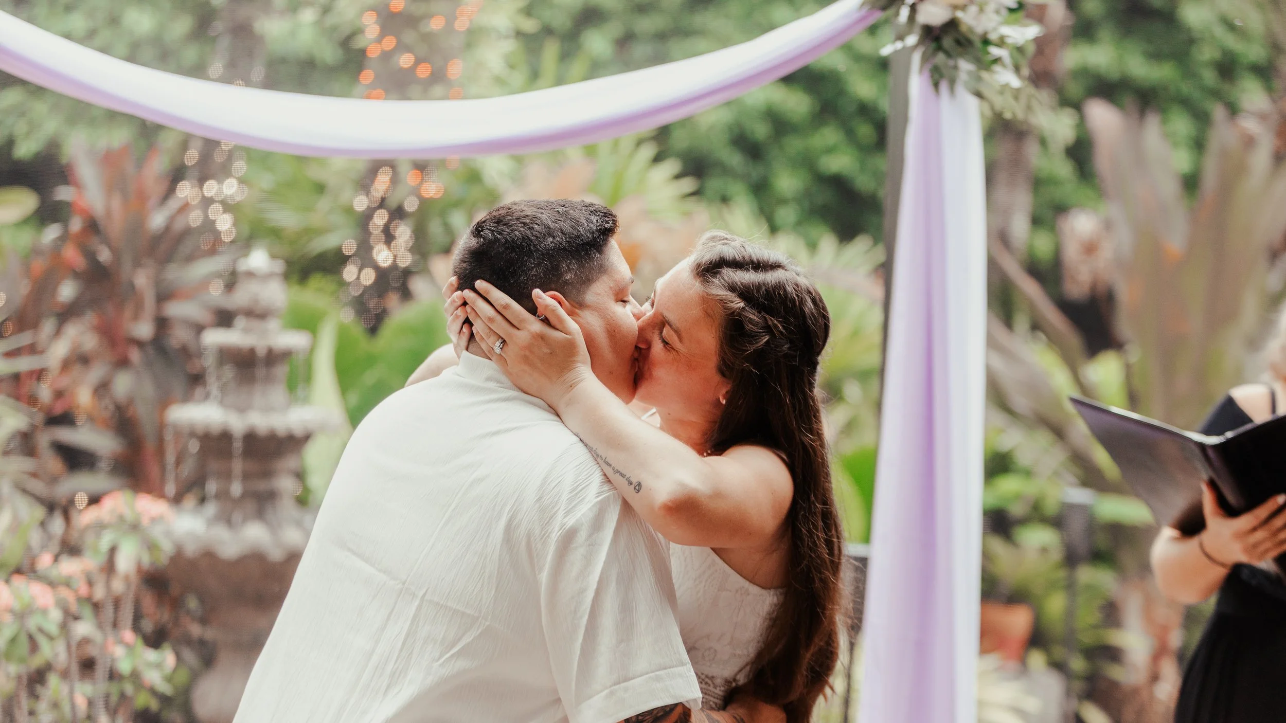 A couple kissing during a wedding ceremony outdoors, with a wedding officiant holding a book nearby and decorative drapery and greenery in the background.