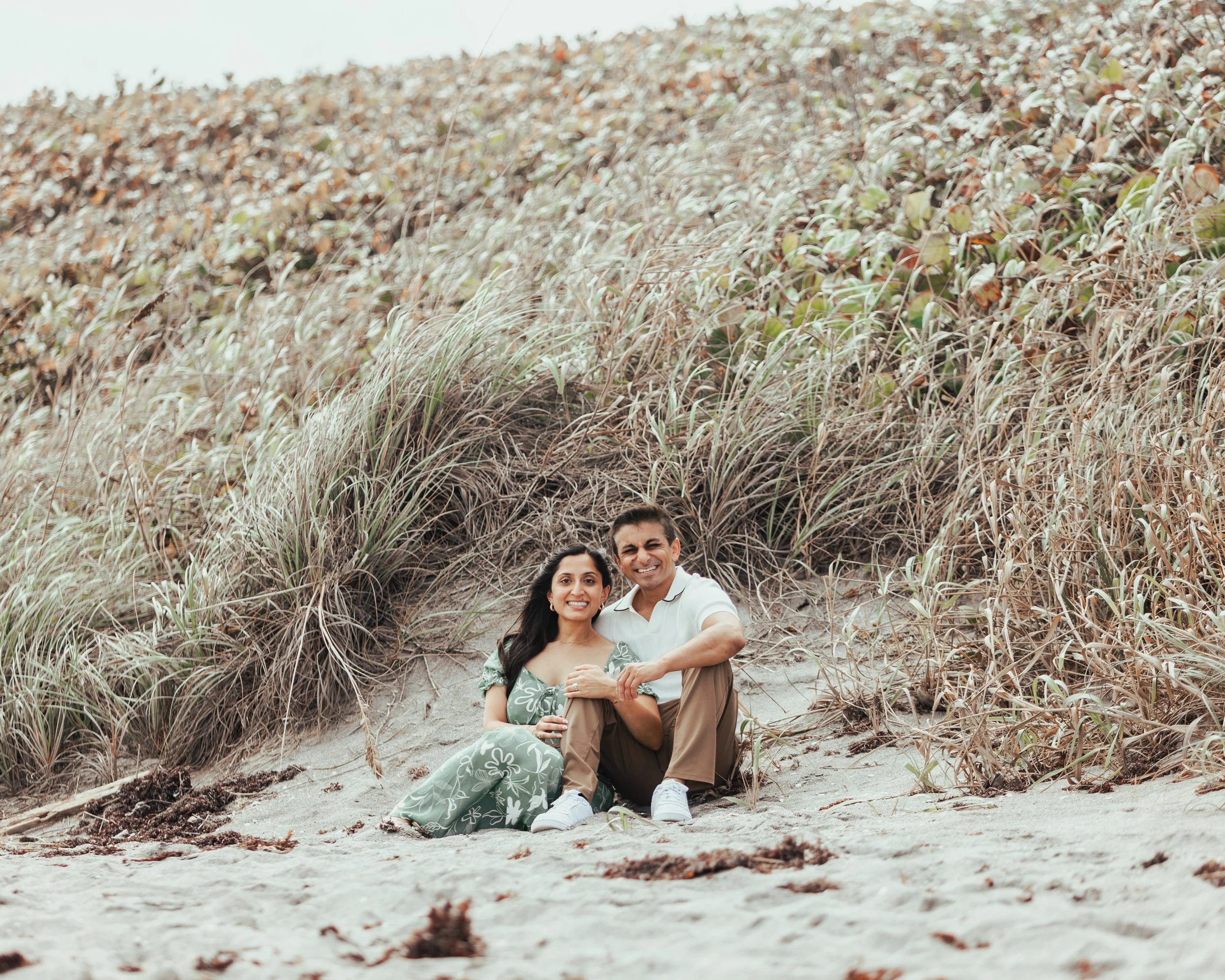 Smiling couple sitting on the sandy beach in front of dune grass and foliage.