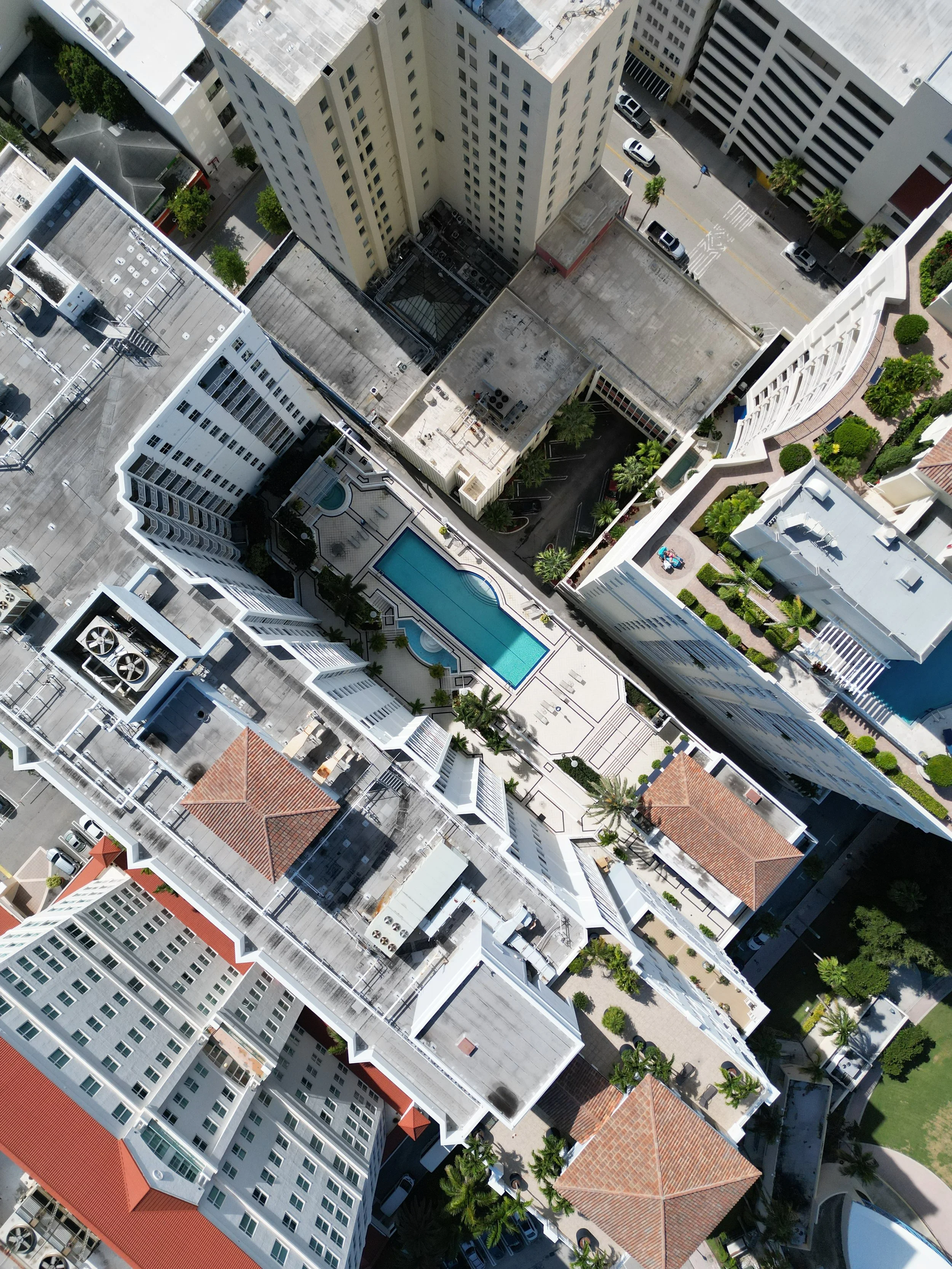 Aerial view of city buildings with swimming pool surrounded by high-rise apartments, trees, and streets with parked cars.