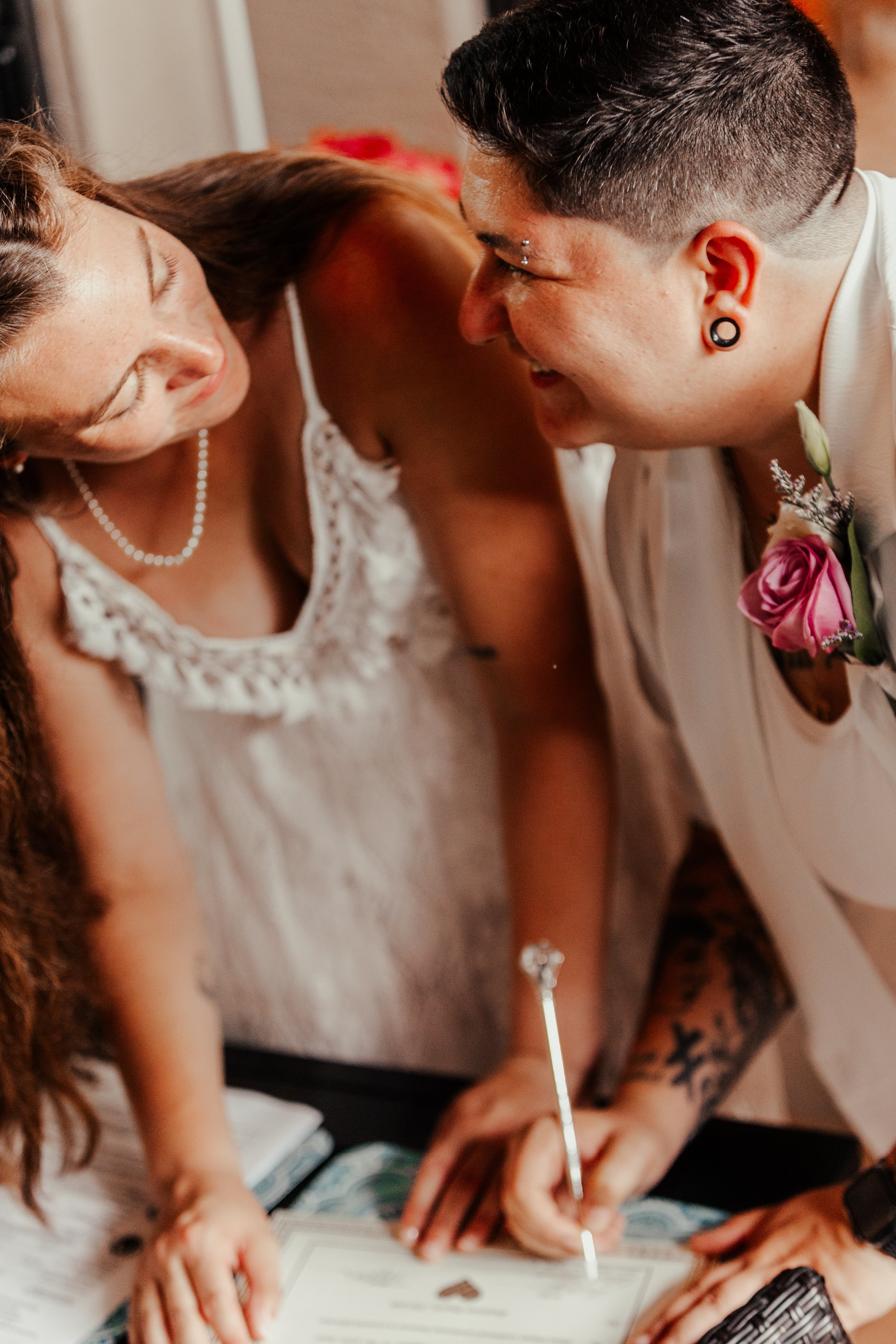 A same-sex couple signing a marriage certificate during their wedding ceremony, with one person holding a pen and both leaning close together.