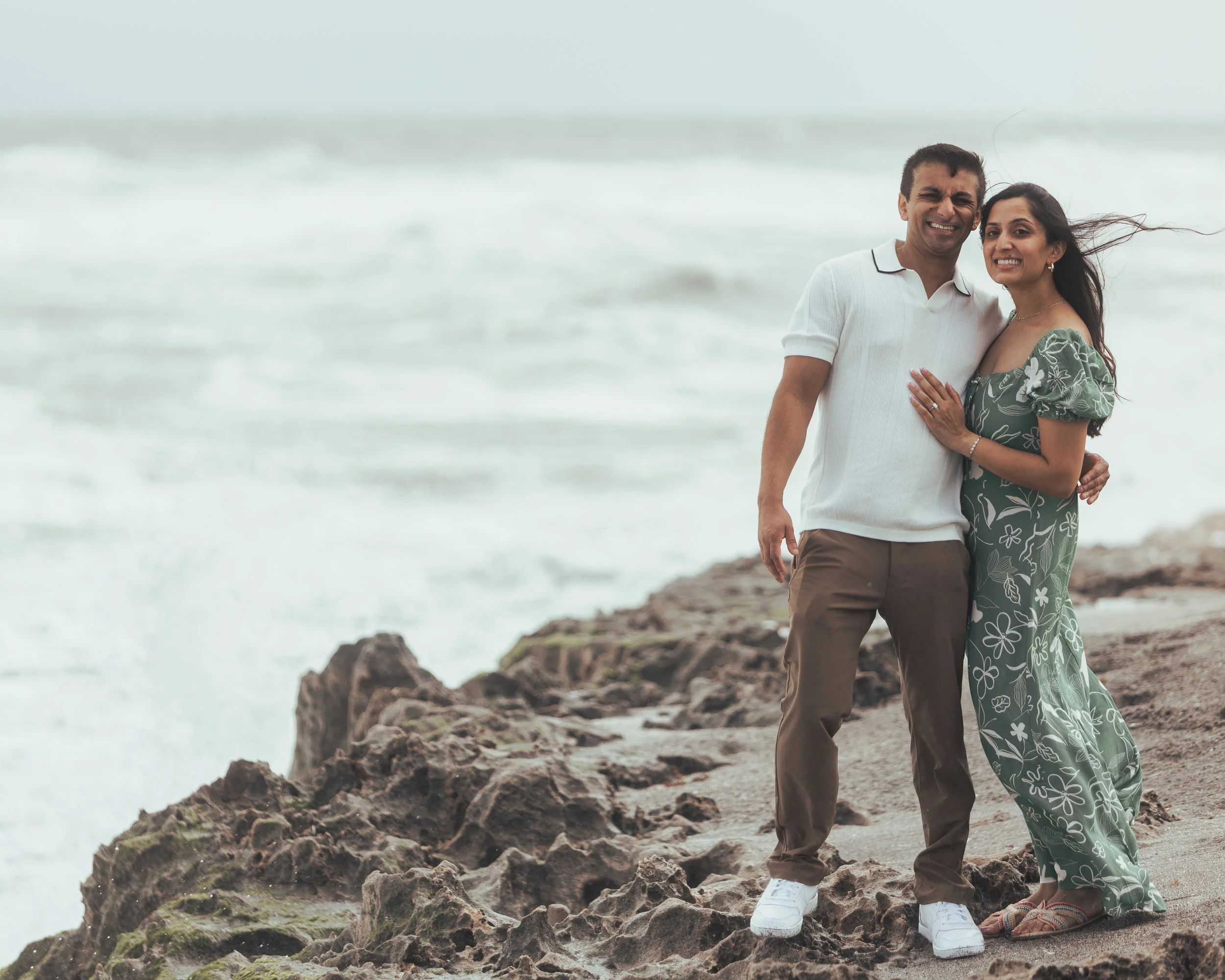 A smiling couple standing on a rocky beach with the ocean in the background, embracing each other.