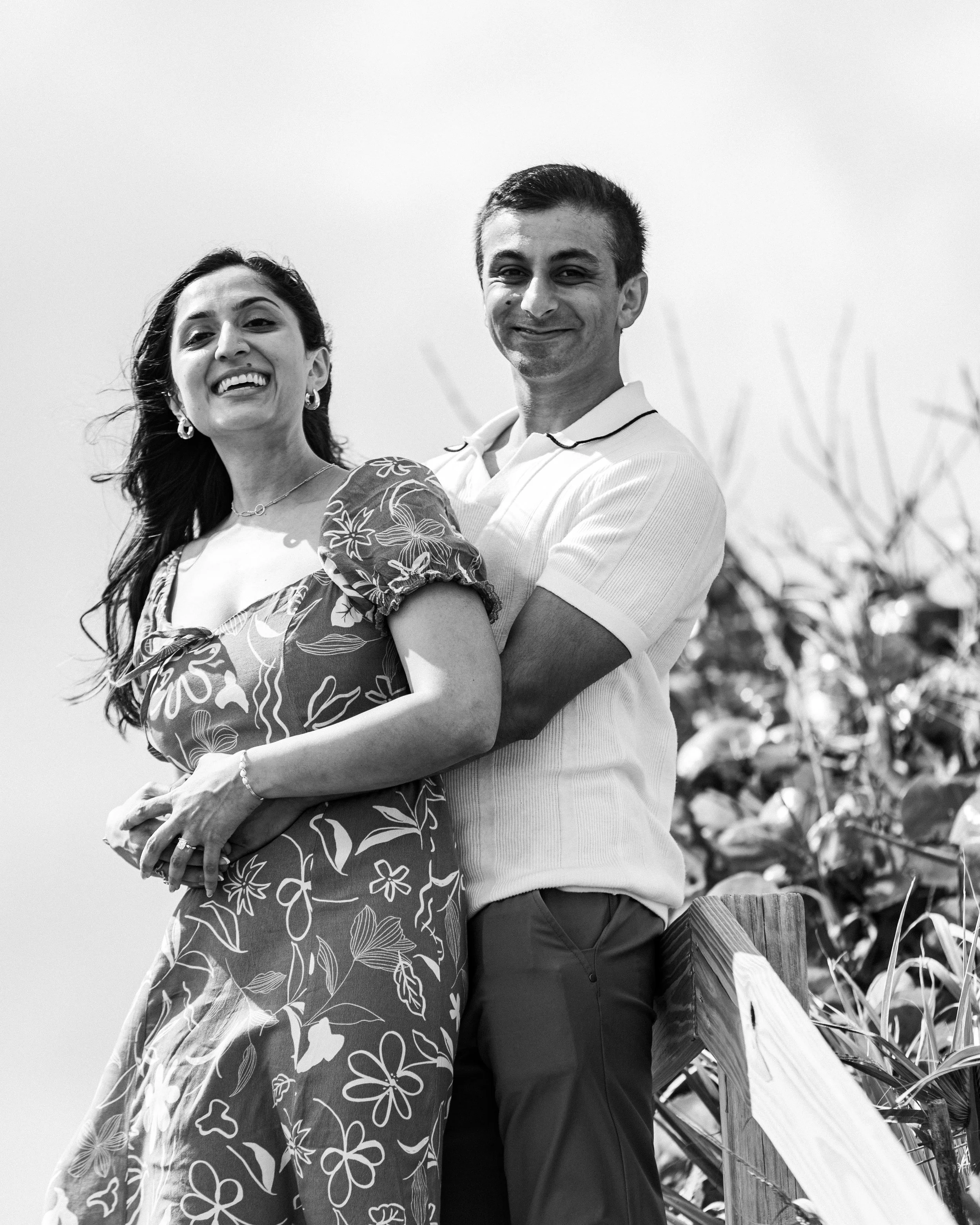 A smiling couple standing outdoors, with the woman wearing a floral dress and the man in a white polo shirt, near a fence and a farm field.