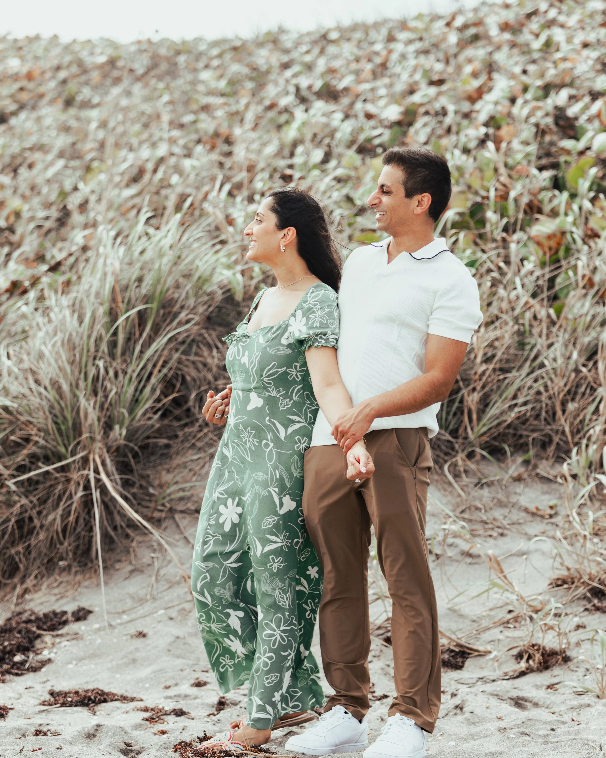 A smiling couple holding hands and walking on a sandy trail near dunes and tall grasses.