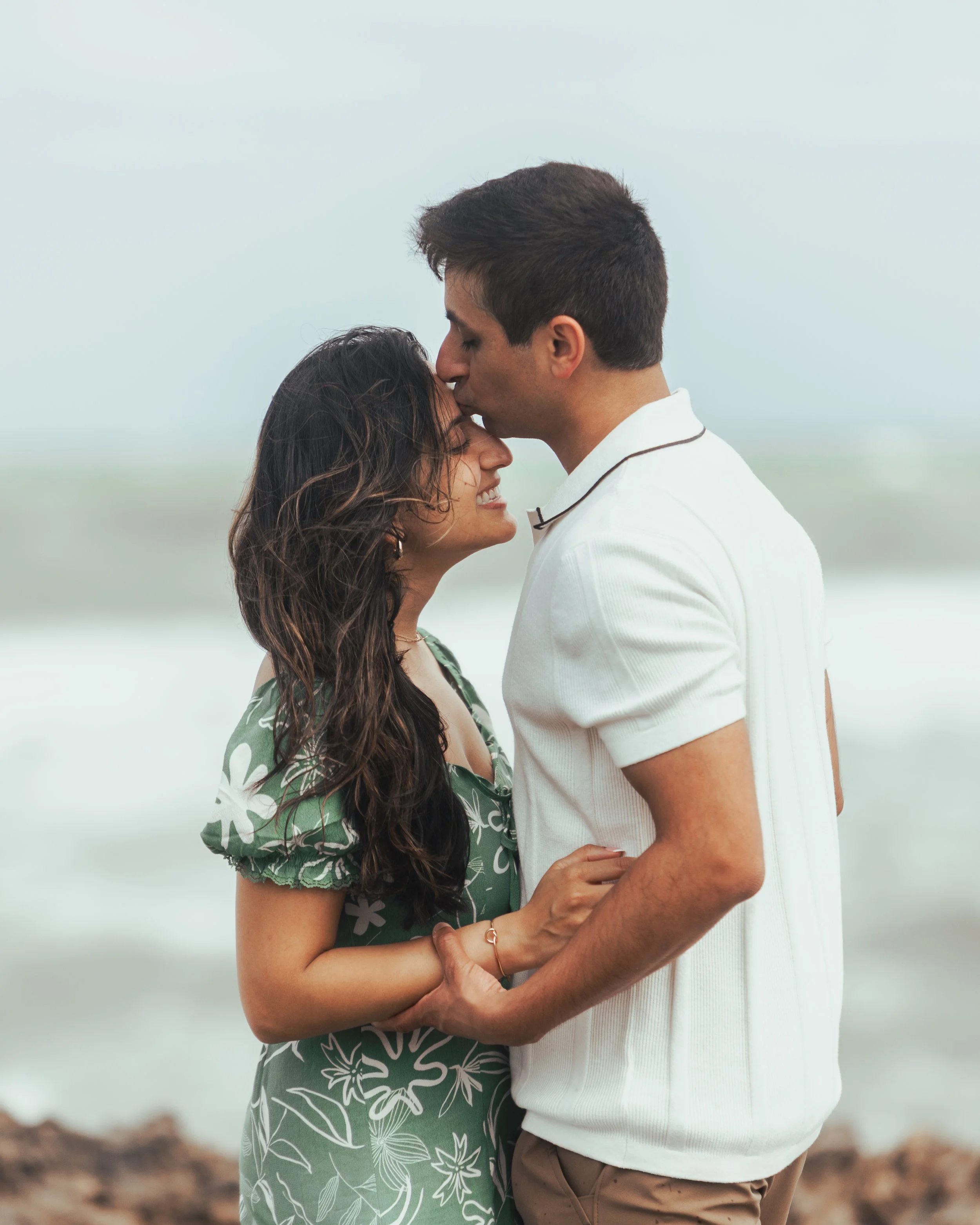 A loving couple standing on the beach, with the man kissing the woman's forehead as they hold hands and smile.