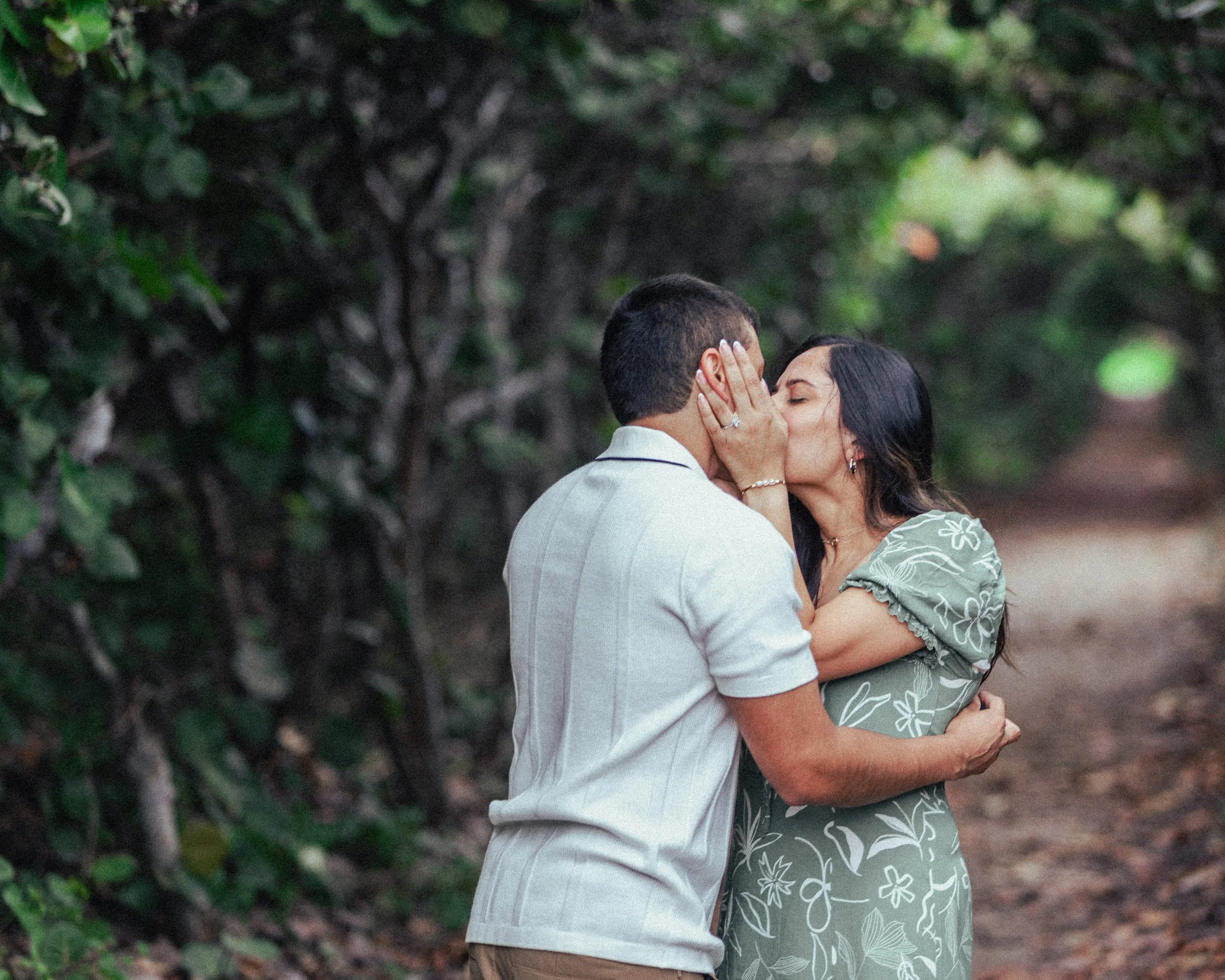 A couple sharing a kiss on a forest trail with green foliage surrounding them.