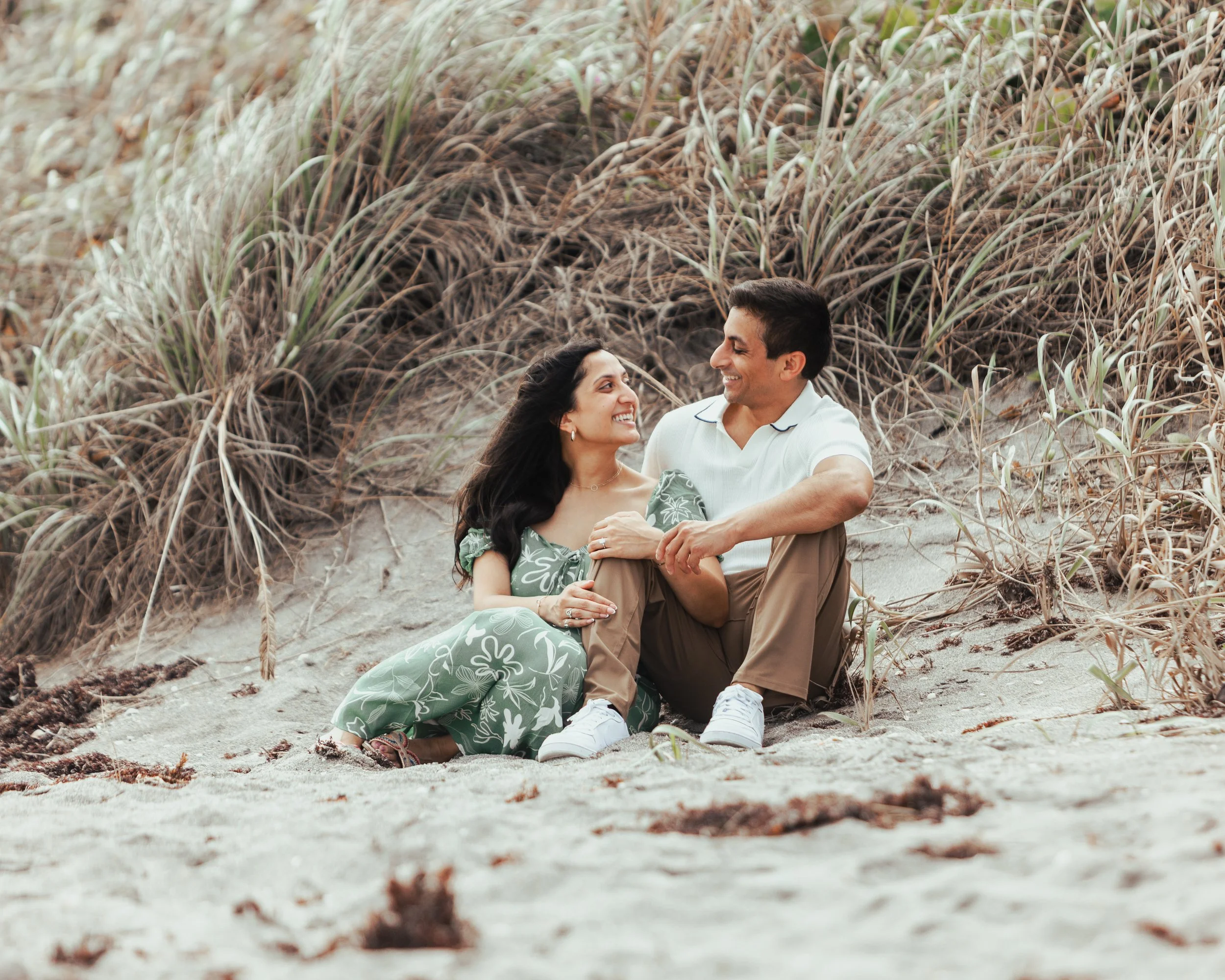 A couple sitting on the sandy beach surrounded by beach grass, smiling and looking at each other.