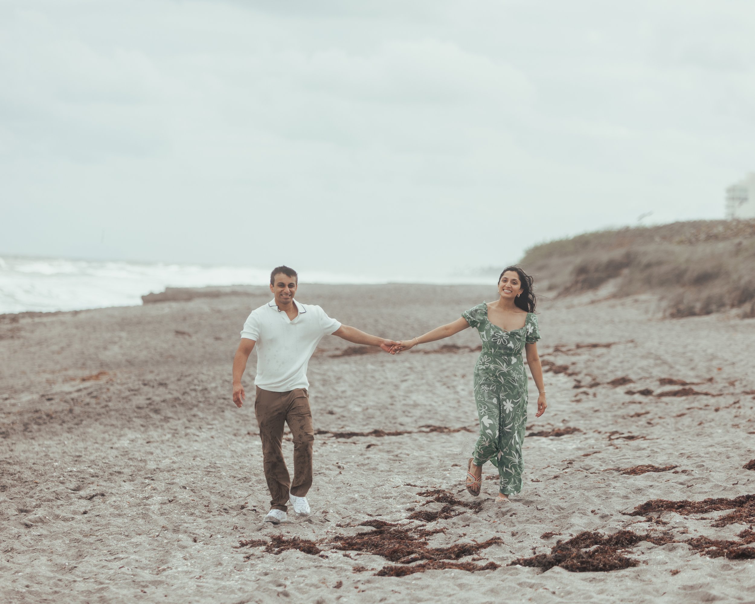 A smiling couple holding hands and walking on a sandy beach, with ocean waves and a cloudy sky in the background.