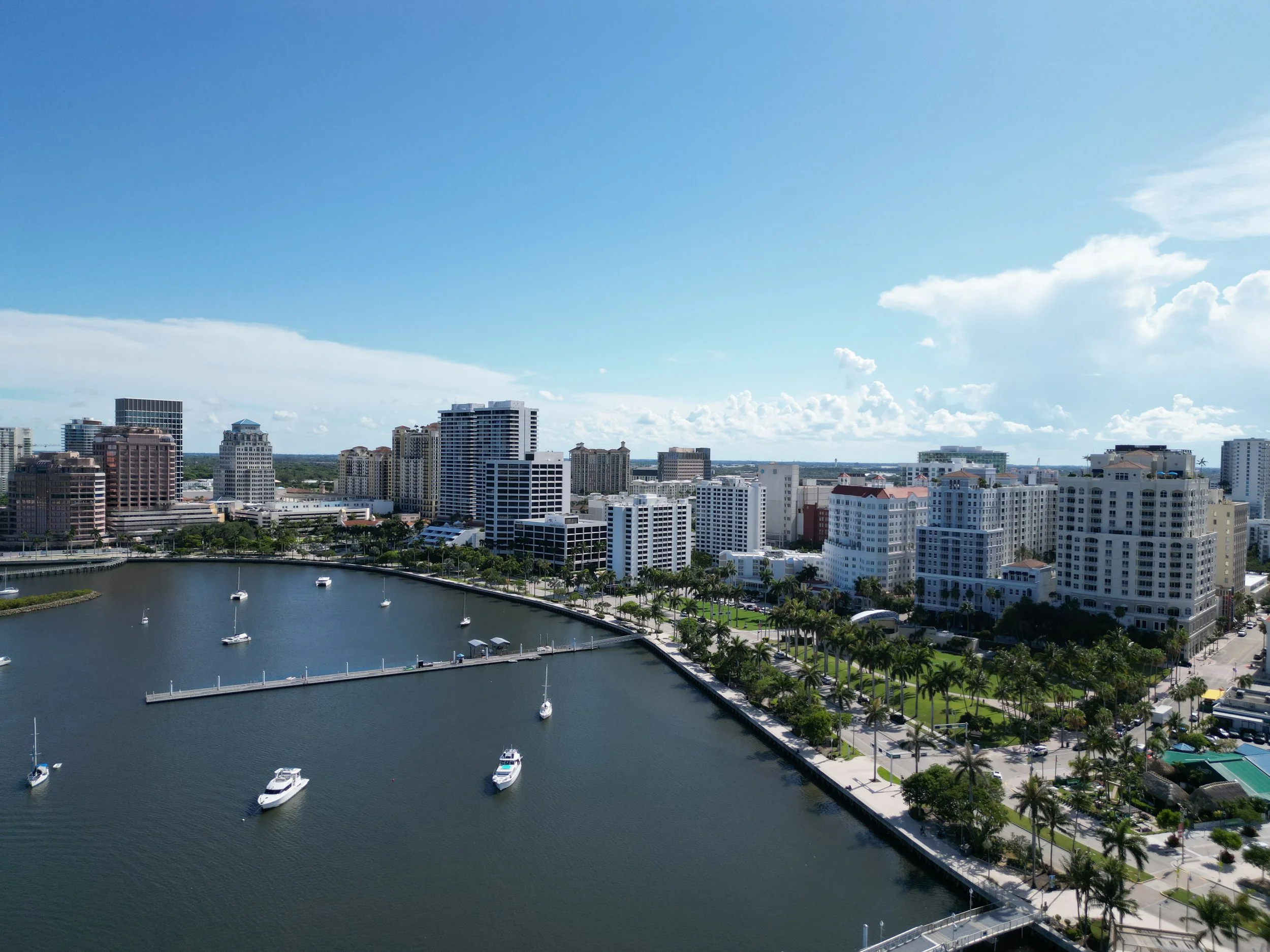 Aerial view of a city skyline with high-rise buildings and a marina with boats on the water, under a partly cloudy sky.