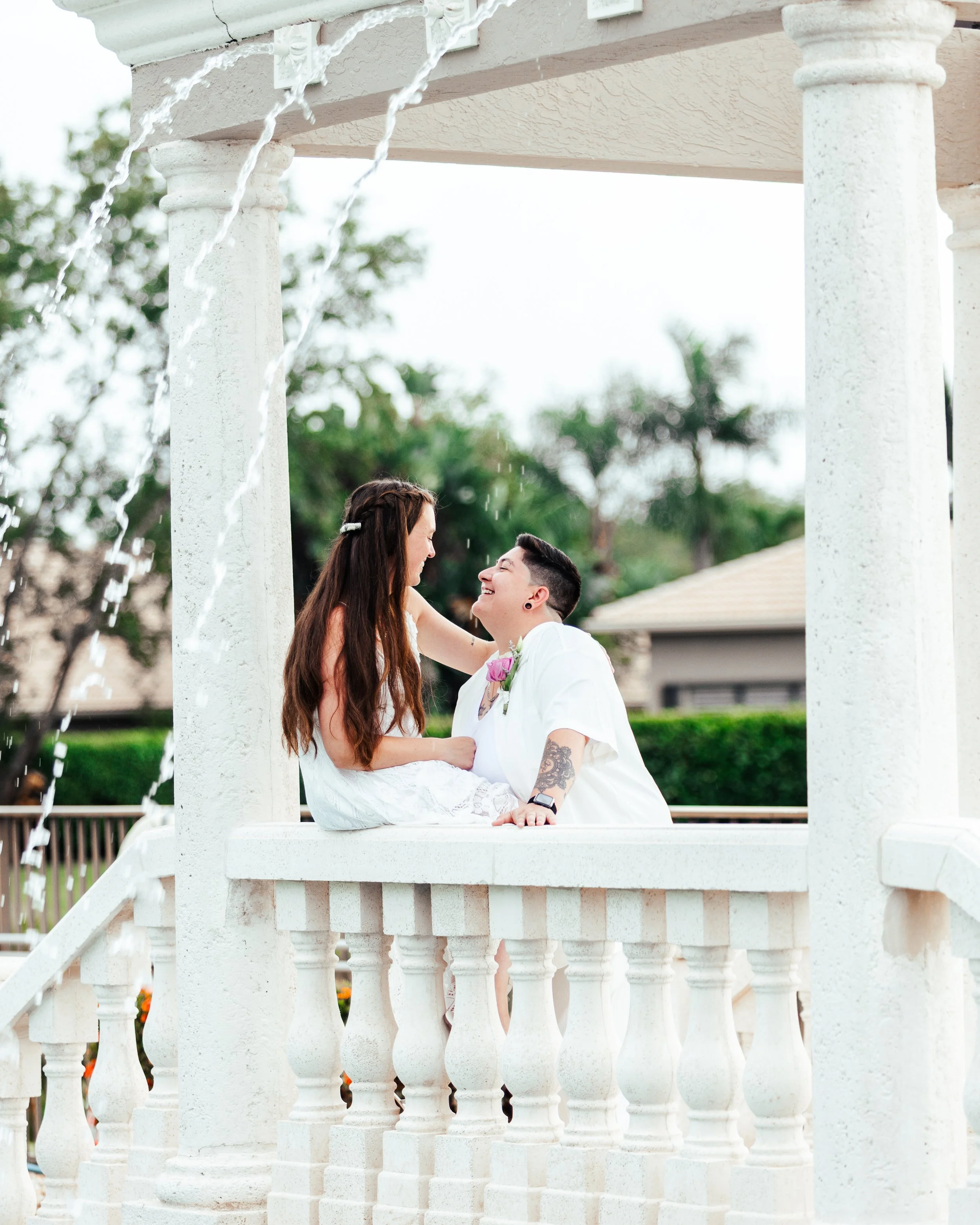 A couple with rainbow flags in their hair sharing a joyful moment on a white gazebo with water features, outdoors on a bright day.