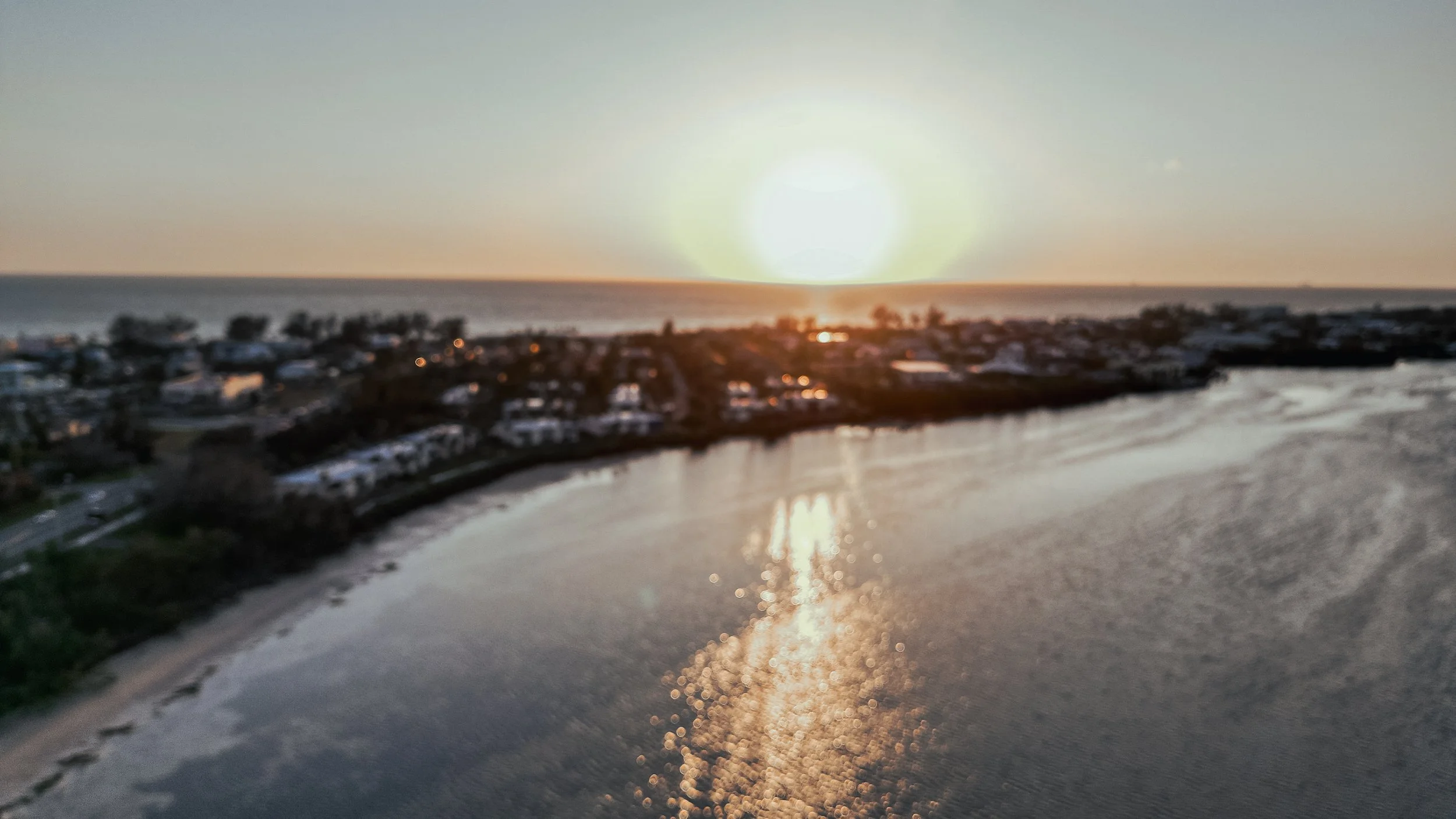 Sun setting over a beach town with buildings, water, and a shoreline in the foreground, with reflective light on the water.