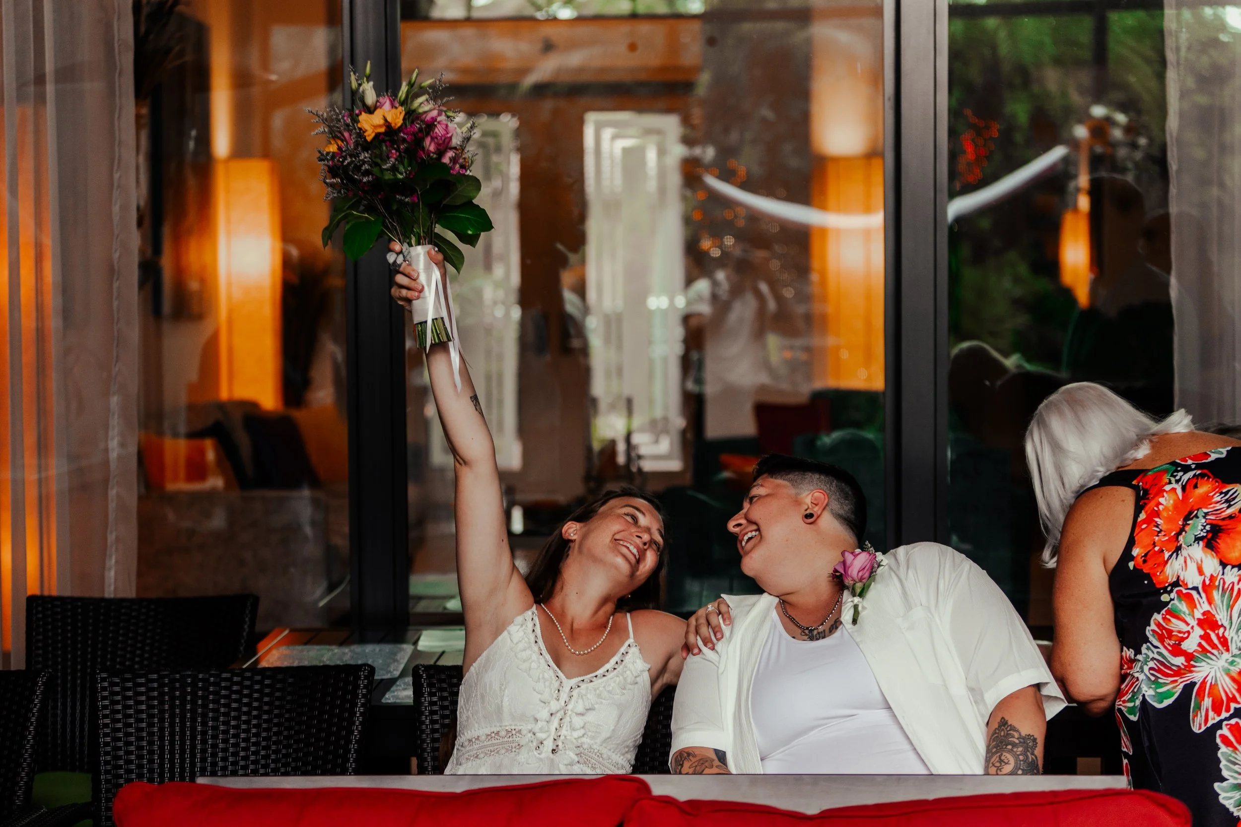 A woman in a white dress holding a bouquet of flowers overhead, smiling at a person with short hair and tattoos, sitting beside her, both smiling and looking at each other.