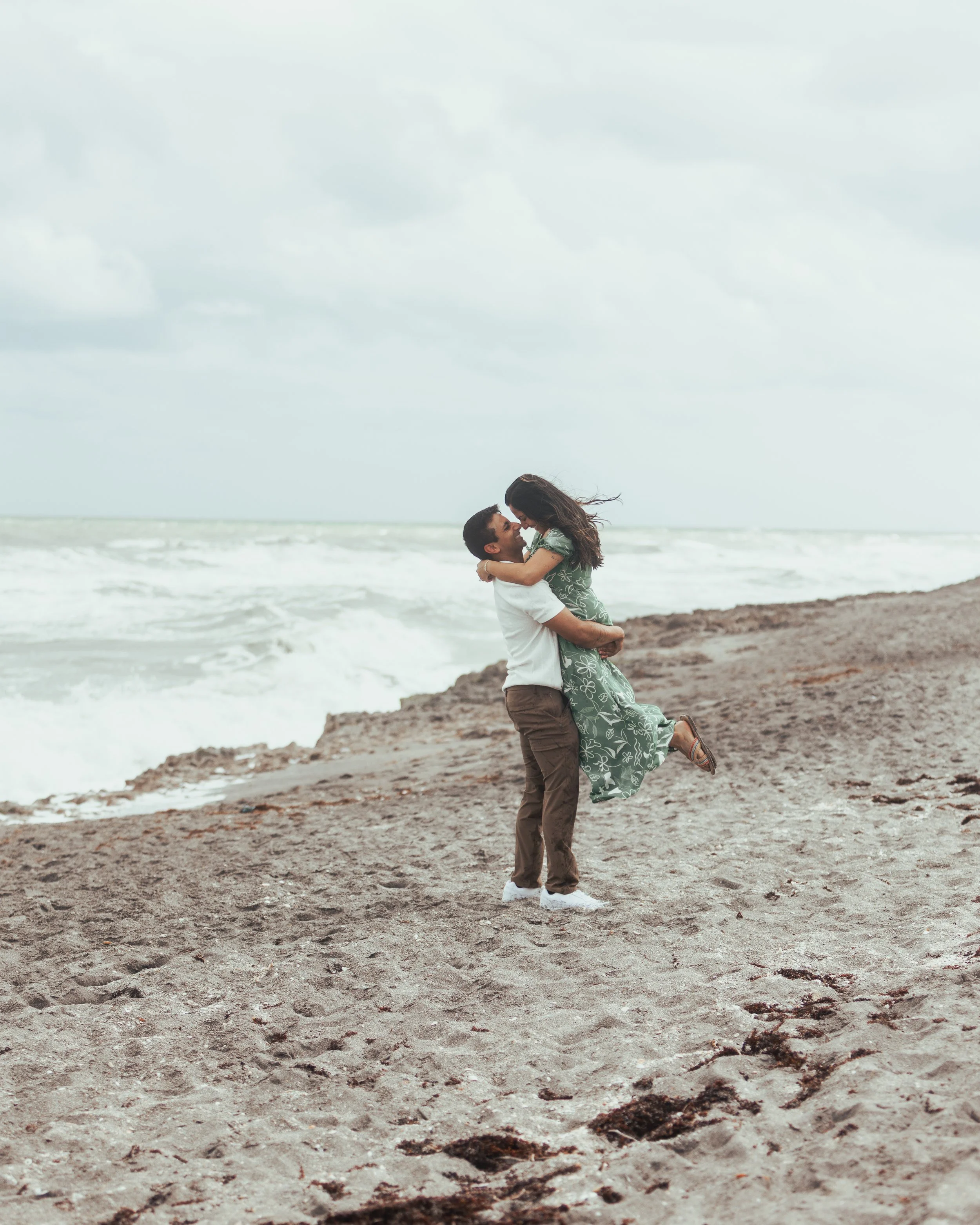 A couple on the beach, the man lifting the woman in his arms as they look at each other, with ocean waves and cloudy sky in the background.