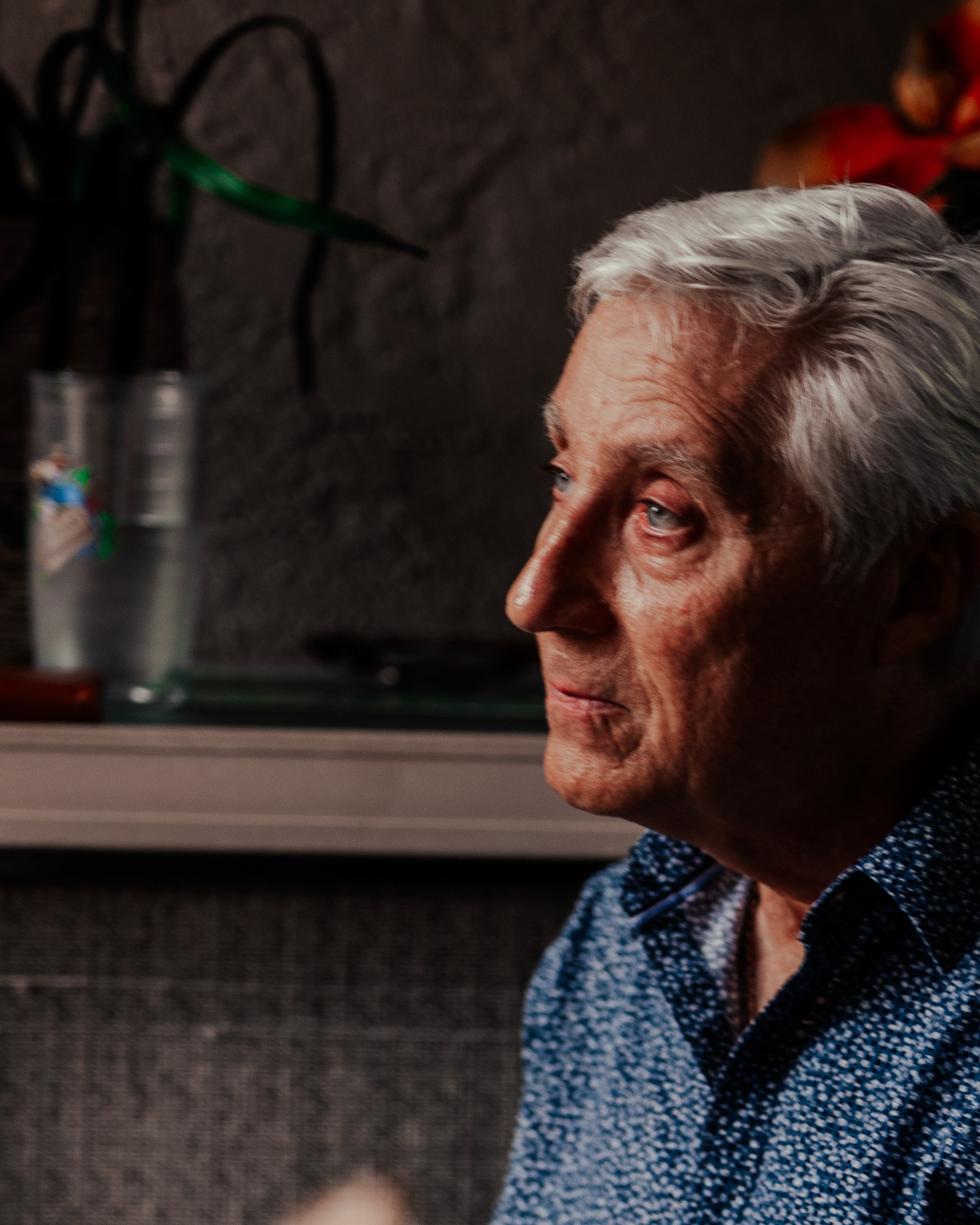 Profile of an elderly man with gray hair, wearing a blue patterned shirt, looking thoughtfully to the right, with a dark background and blurred objects behind him.