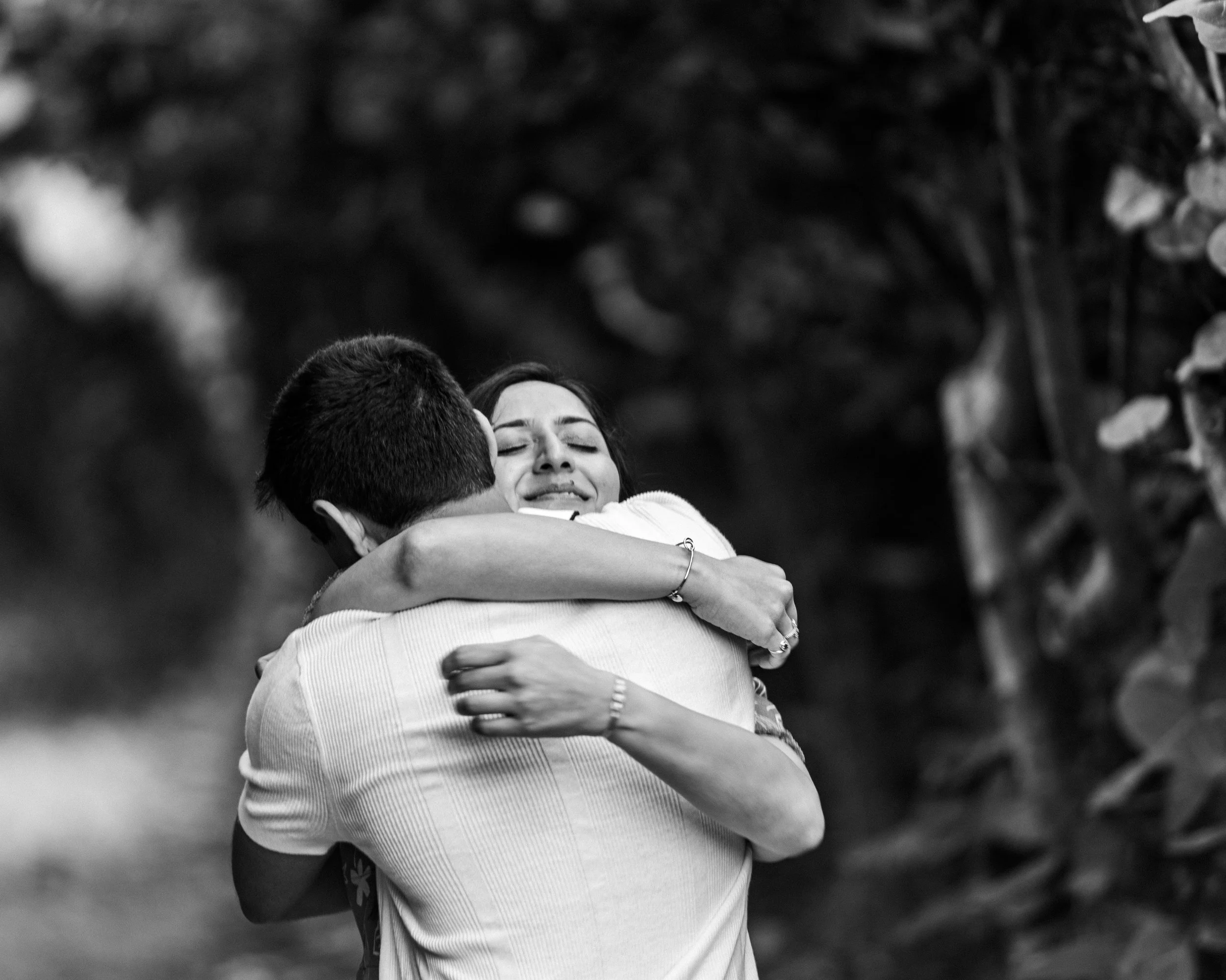A woman and a man hug each other tightly, with the woman's eyes closed and a look of emotion on her face, as they embrace outdoors.