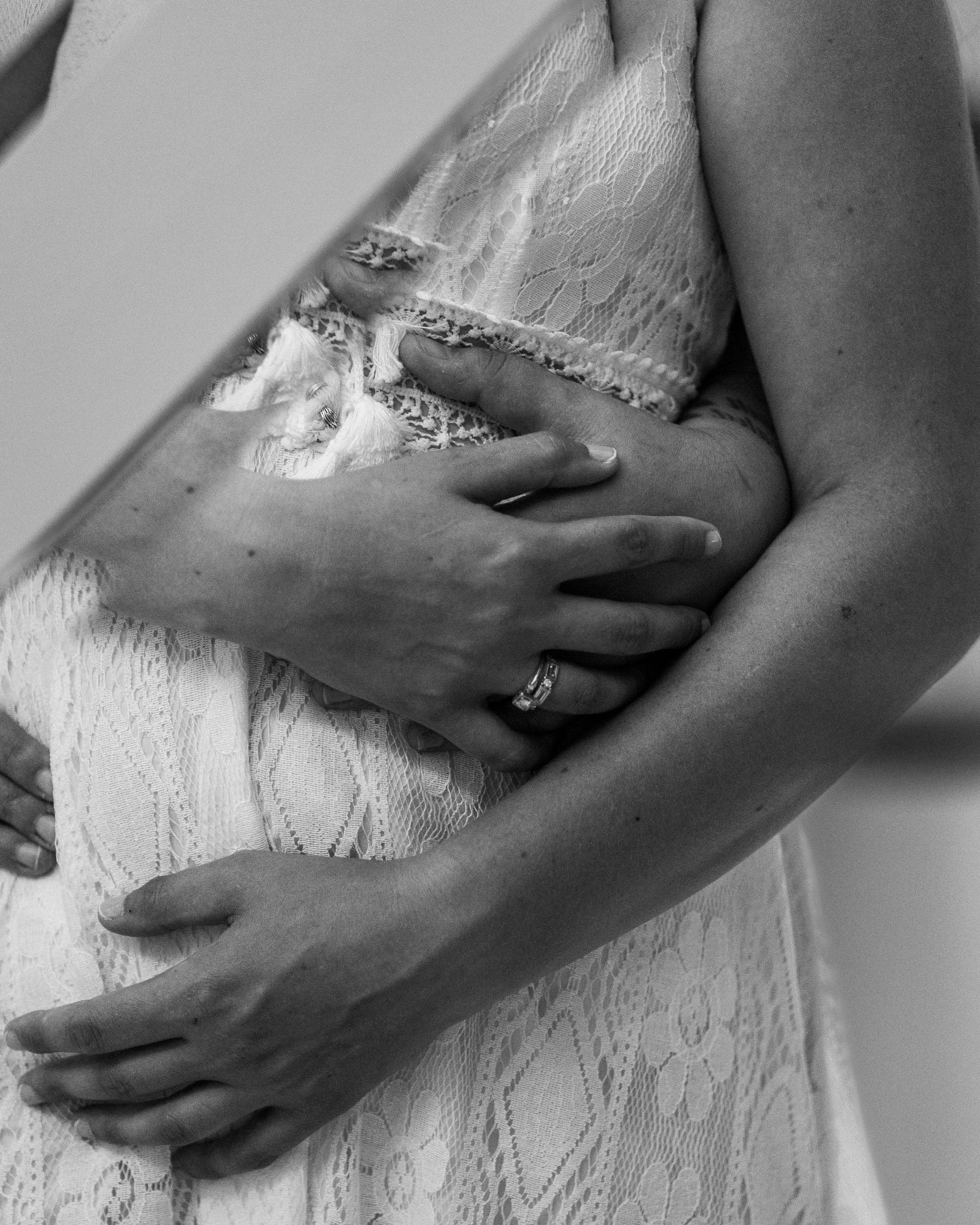 A woman wearing a lace dress holding her pregnant belly, with her hands gently resting on it, and wearing a wedding ring.