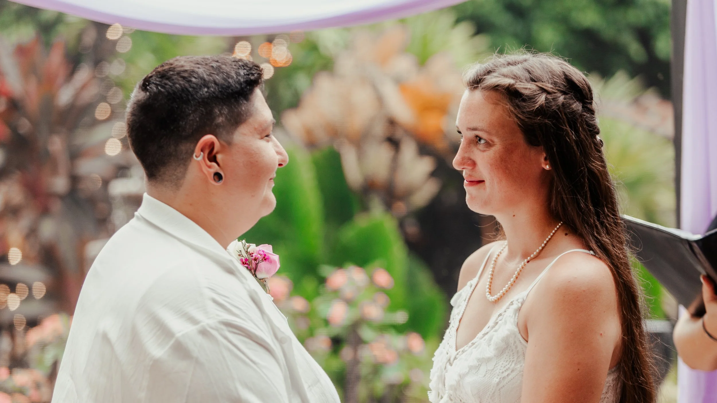 A same-sex couple standing close together during their wedding ceremony outdoors, smiling at each other with blurred trees and flowers in the background.