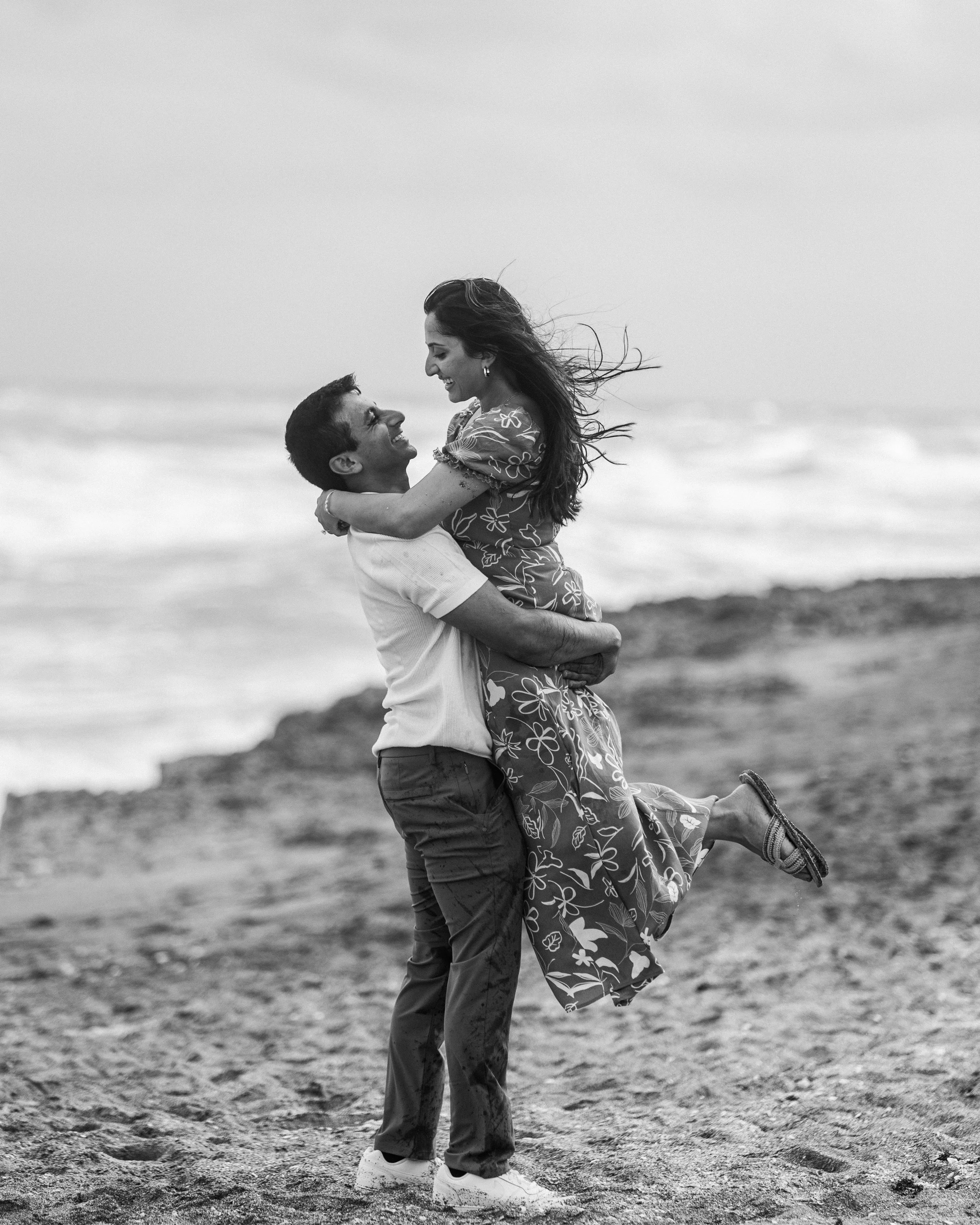 A joyful couple on the beach with the man lifting the woman in his arms, both smiling and looking at each other.