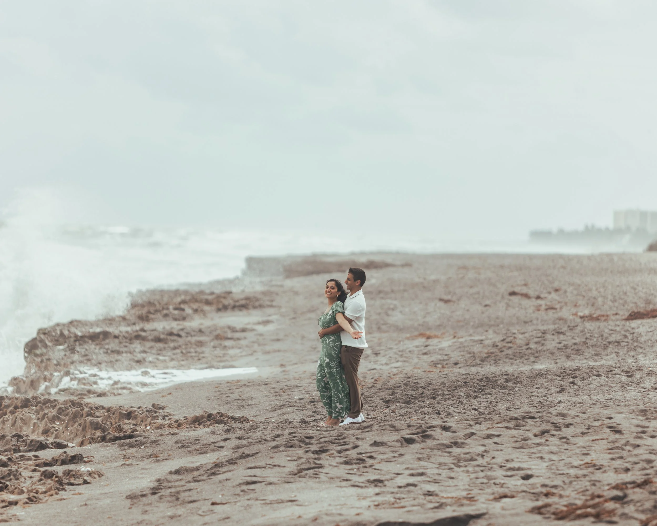A couple standing on a sandy beach near the ocean, hugging and smiling, with waves crashing behind them under a cloudy sky.