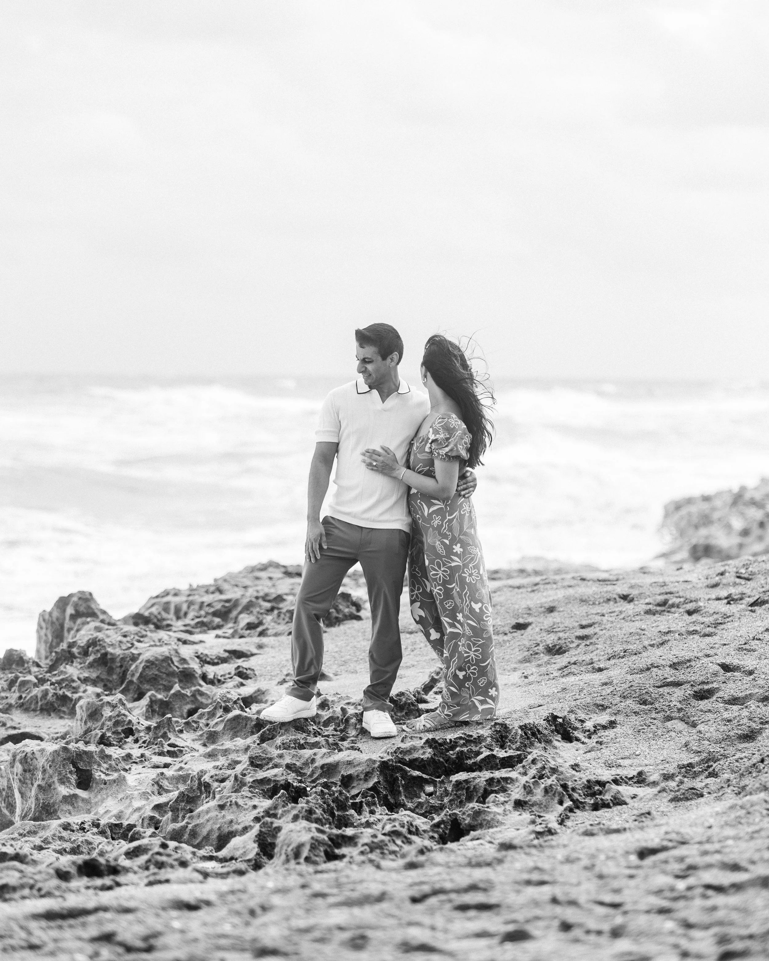 A young couple standing on rocks at the beach, smiling and embracing each other with waves in the background.